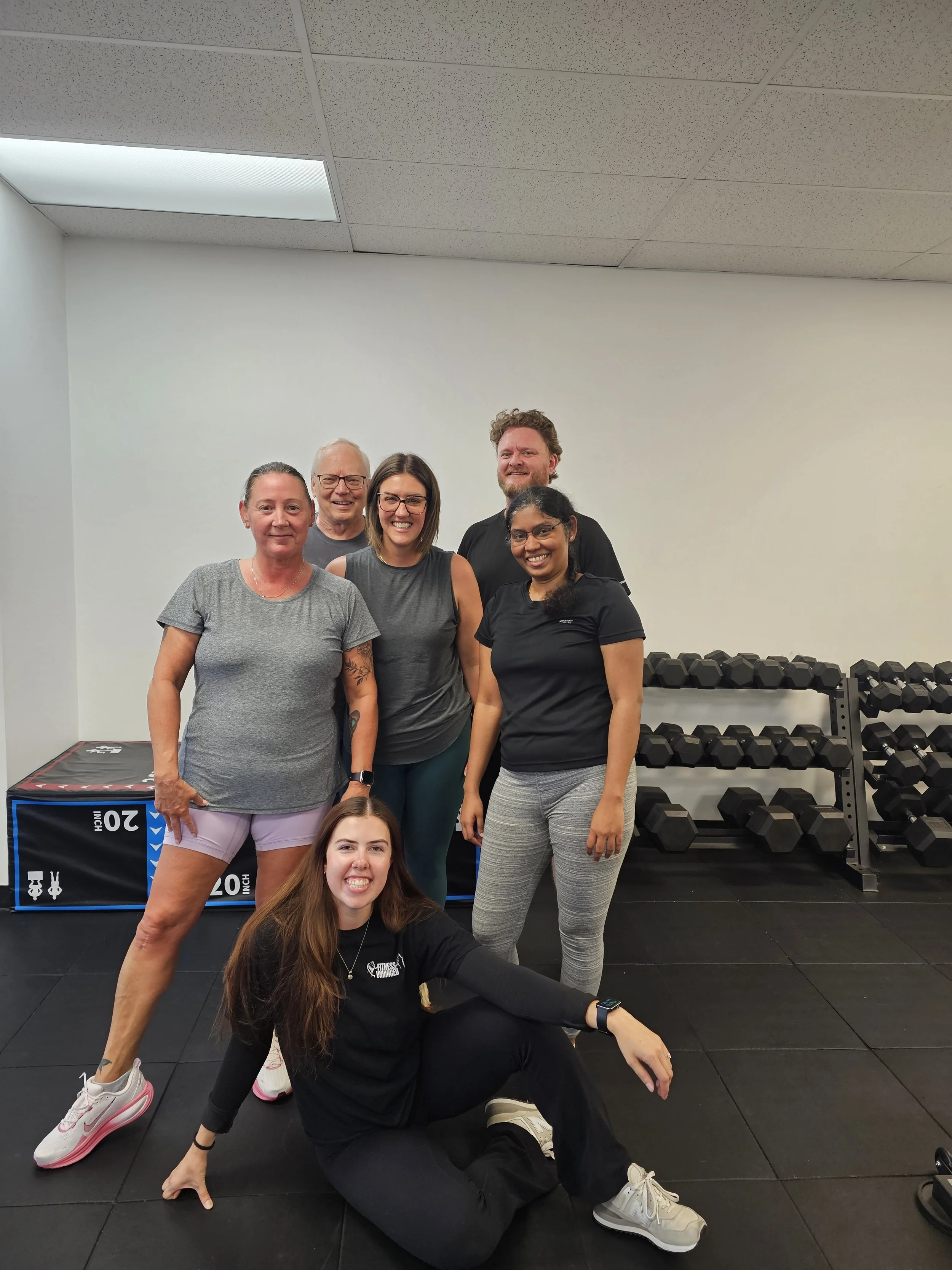 Group of five people at the gym, smiling and posing for a photo with weightlifting equipment and a black floor.