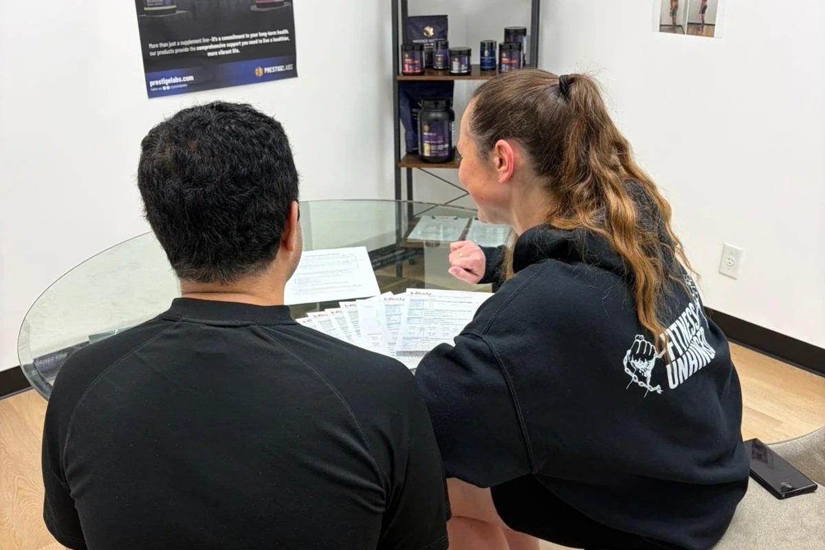 A woman and a man sitting at a glass table, engaged in a conversation, with papers spread out on the table. The woman is smiling, wearing a black hoodie with a logo on the back. The man has short, dark hair and is wearing a black shirt. In the background, there are shelves with supplements and a white wall with a poster.