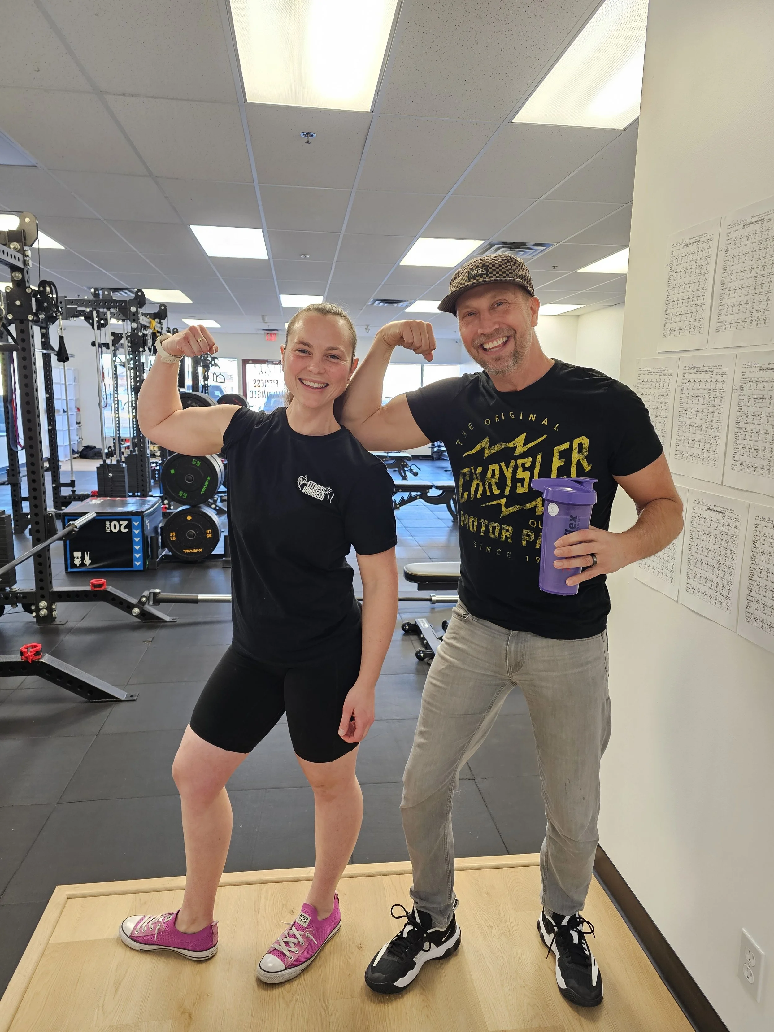 A smiling woman in pink sneakers and a black workout outfit flexing her her arm muscle next to a smiling man in black and yellow t-shirt and khaki pants holding a purple shaker bottle, both in a gym.
