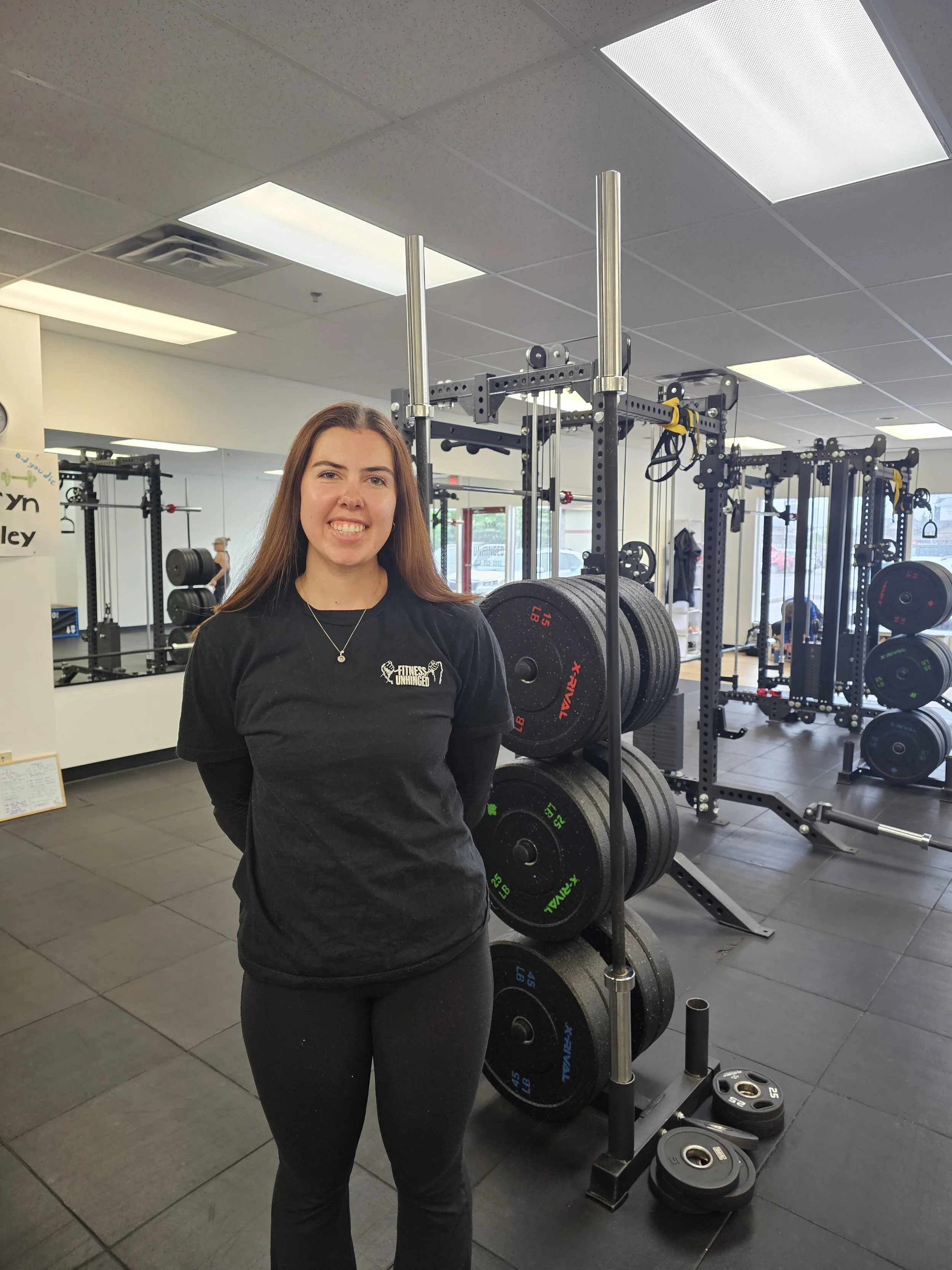 A young woman smiling at the camera inside a gym, standing in front of weightlifting equipment, wearing a black t-shirt with a fitness logo, and black pants.