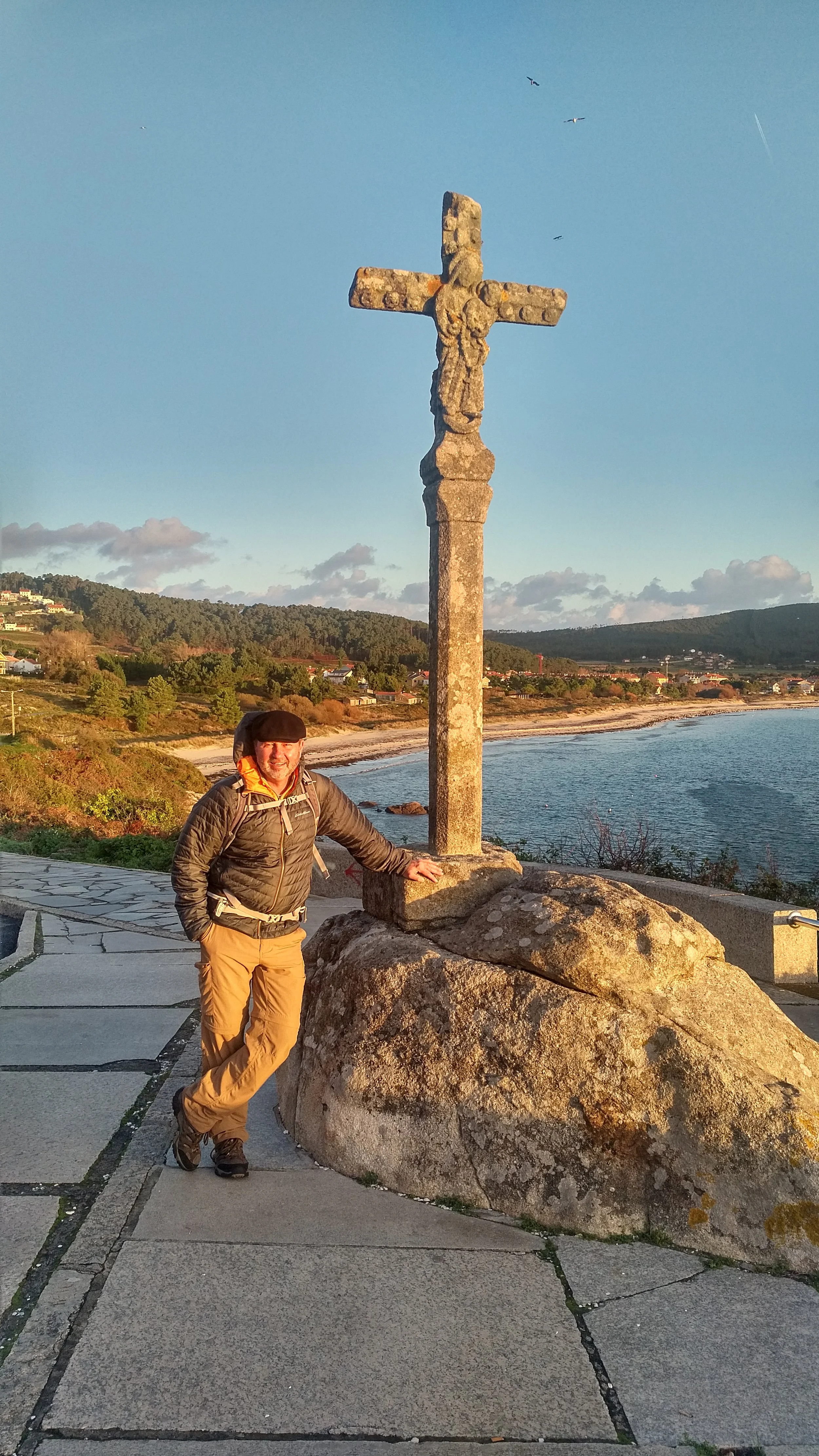 A Camino pilgrim in outdoor clothing with a backpack smiling and posing next to a large stone cross on a coastal walkway with a scenic view of the ocean, hills, and houses in the background during sunrise over Finisterre.