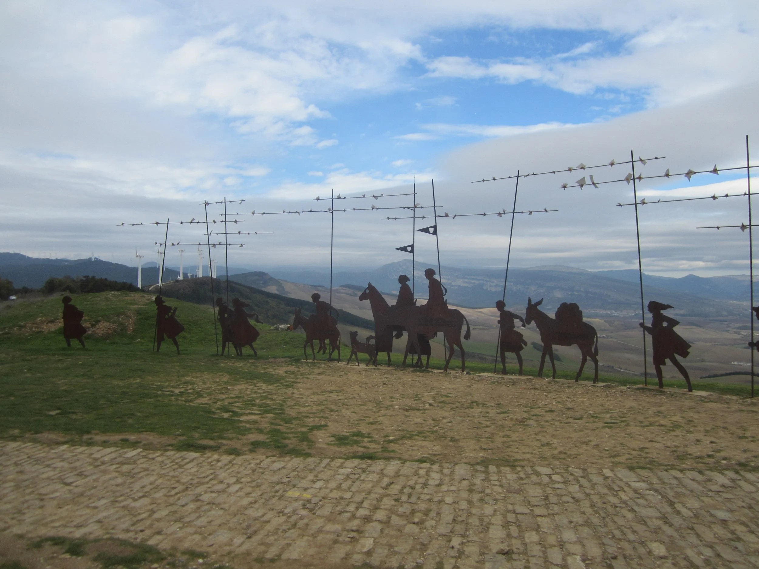 Metal sculptures of cowboys and horses with windmill and wind turbines in the background on a grassy hill under a partly cloudy sky.