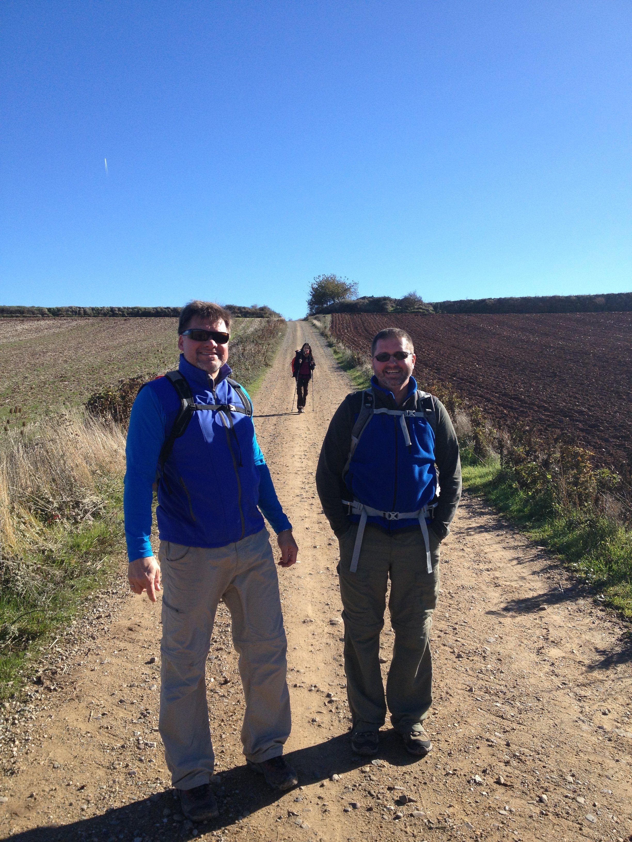 Two men in blue jackets and hiking pants stand on a dirt trail in a rural area, with a woman in a red jacket walking behind them. The landscape features plowed fields, bushes, and small trees under a clear blue sky.