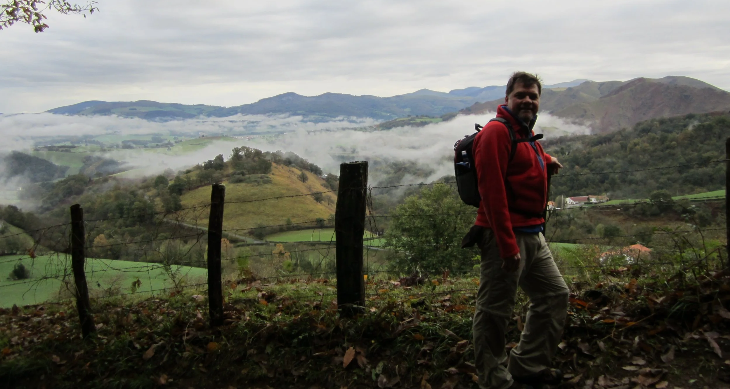 A man hiking outdoors in a hilly, rural landscape with mist and clouds in the background, wearing a red jacket and carrying a backpack.