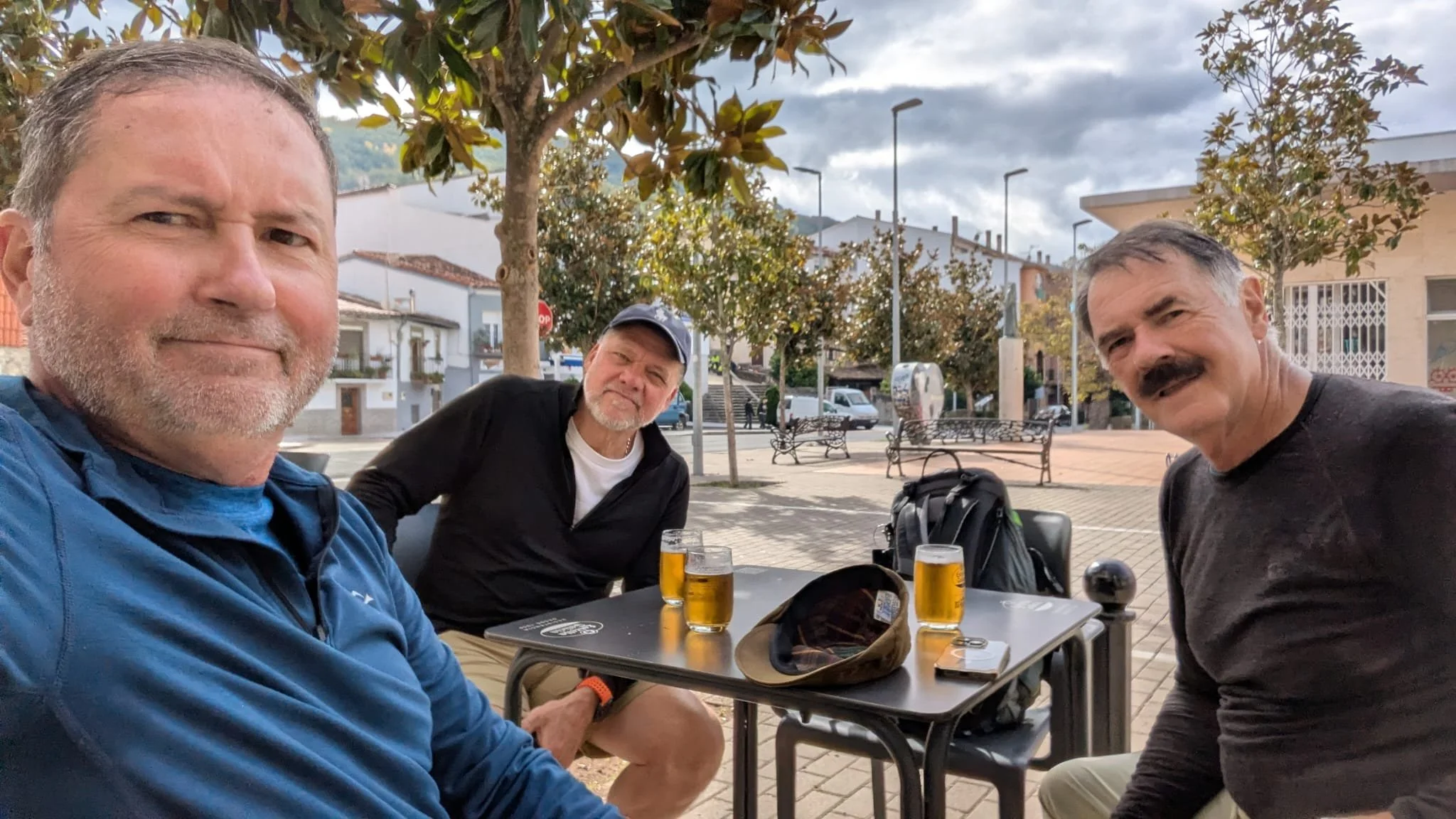 Three middle-aged men sitting at an outdoor table with drinks, surrounded by trees and buildings, enjoying a casual gathering.