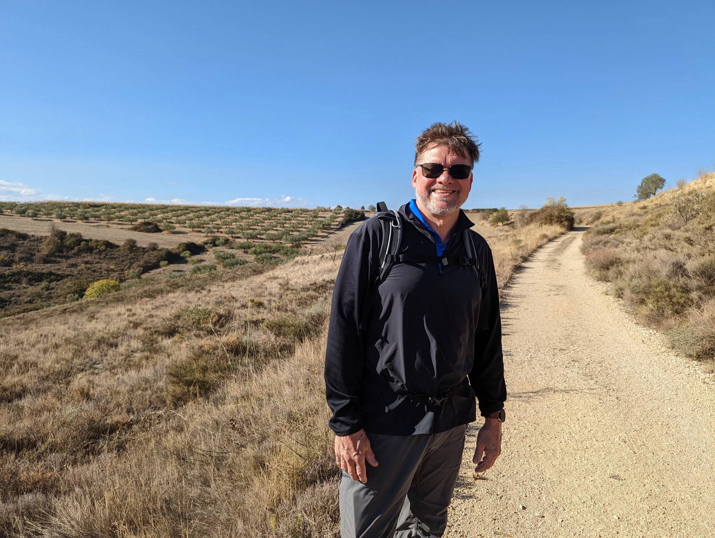 A smiling man wearing sunglasses, a black jacket, and a backpack, standing on a dirt trail in a dry, open landscape with sparse trees and shrubbery, under a clear blue sky.