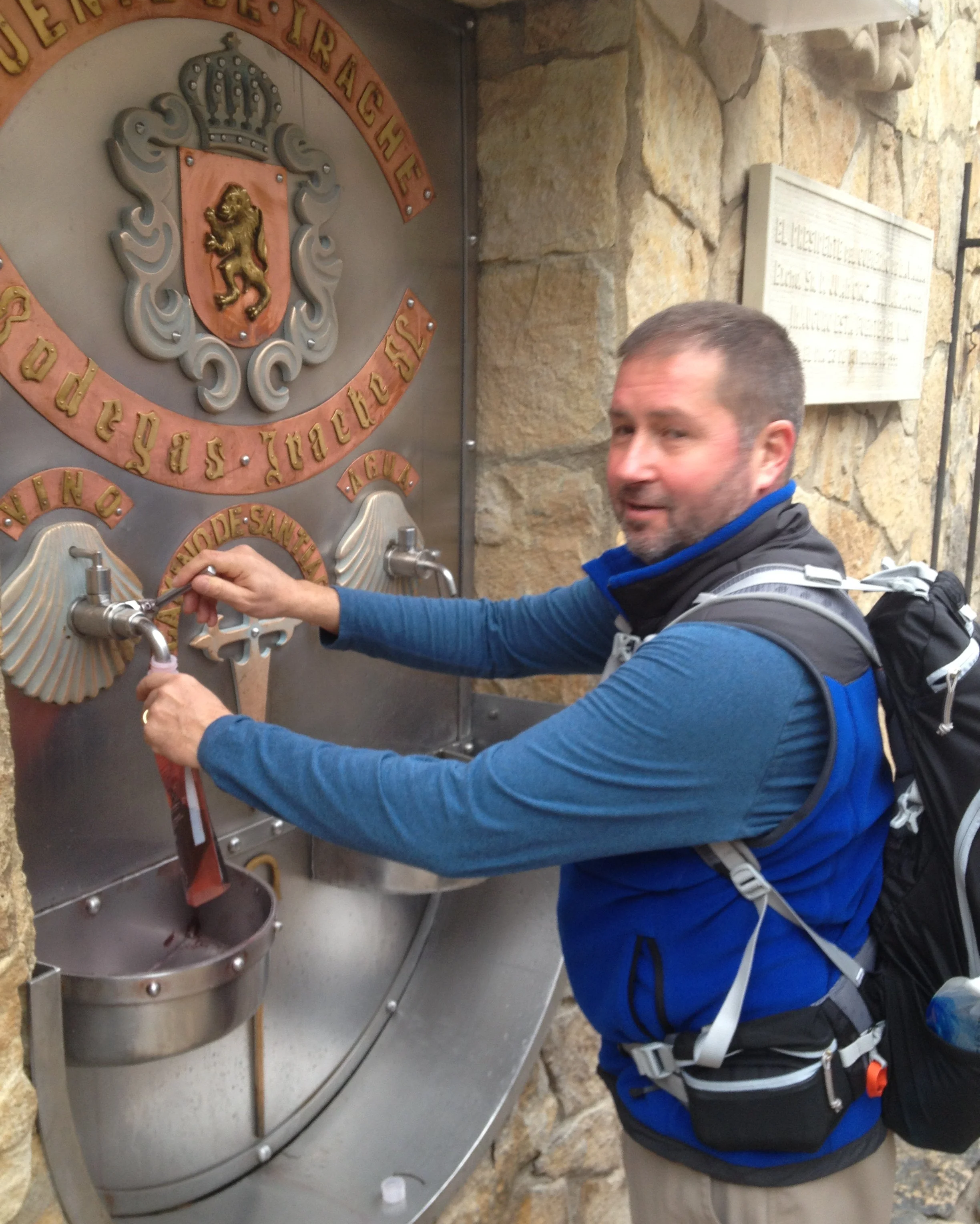 A man in outdoor clothing with a backpack is filling a bottle at a water fountain with a decorative crest on a stone wall behind him.