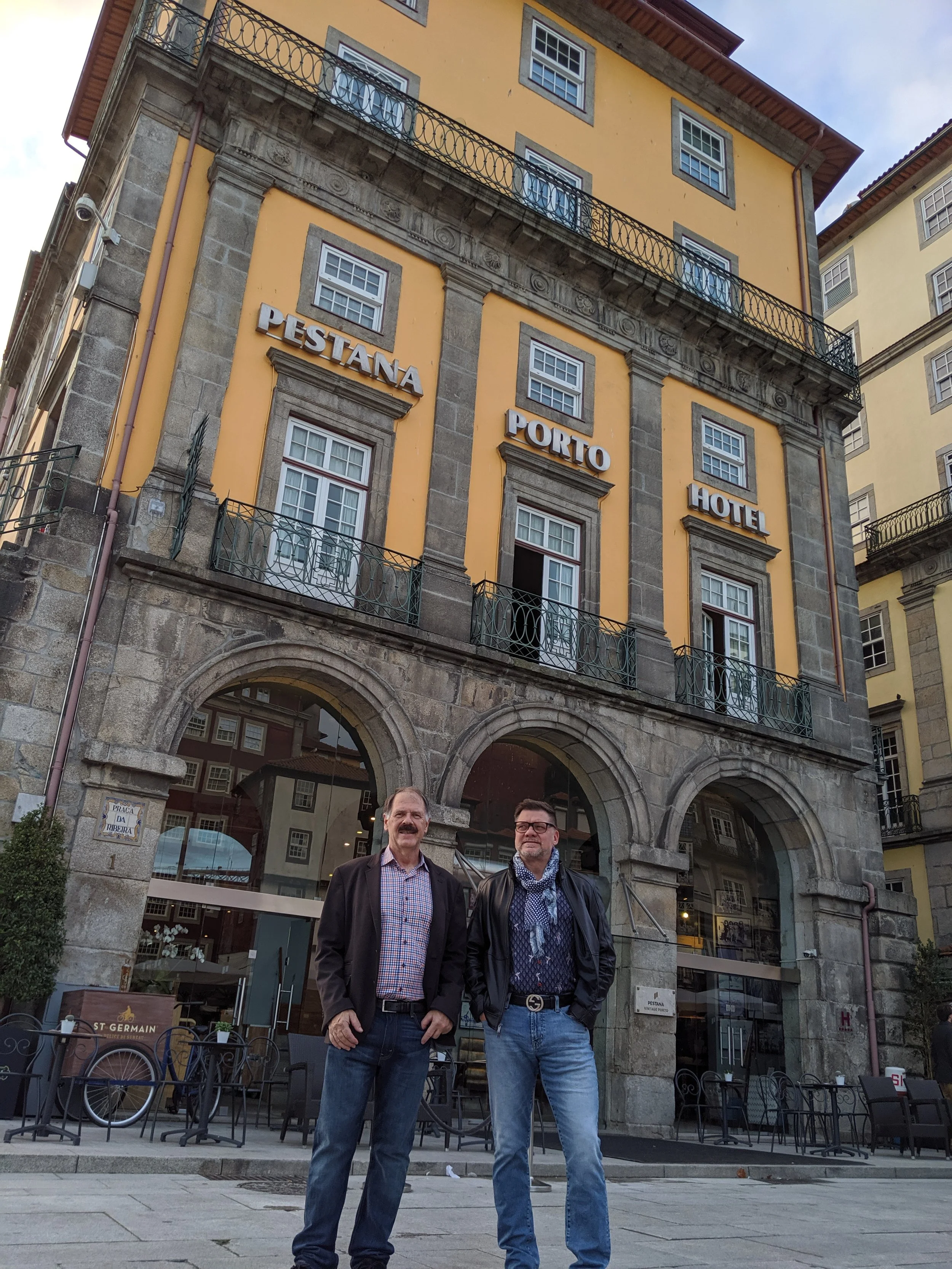 Two men standing outdoors in front of a historic building with yellow walls, large windows, and a sign that reads "Pestana Porto Hotel." They are smiling and dressed in casual business attire. The building has decorative stonework and wrought iron balconies.