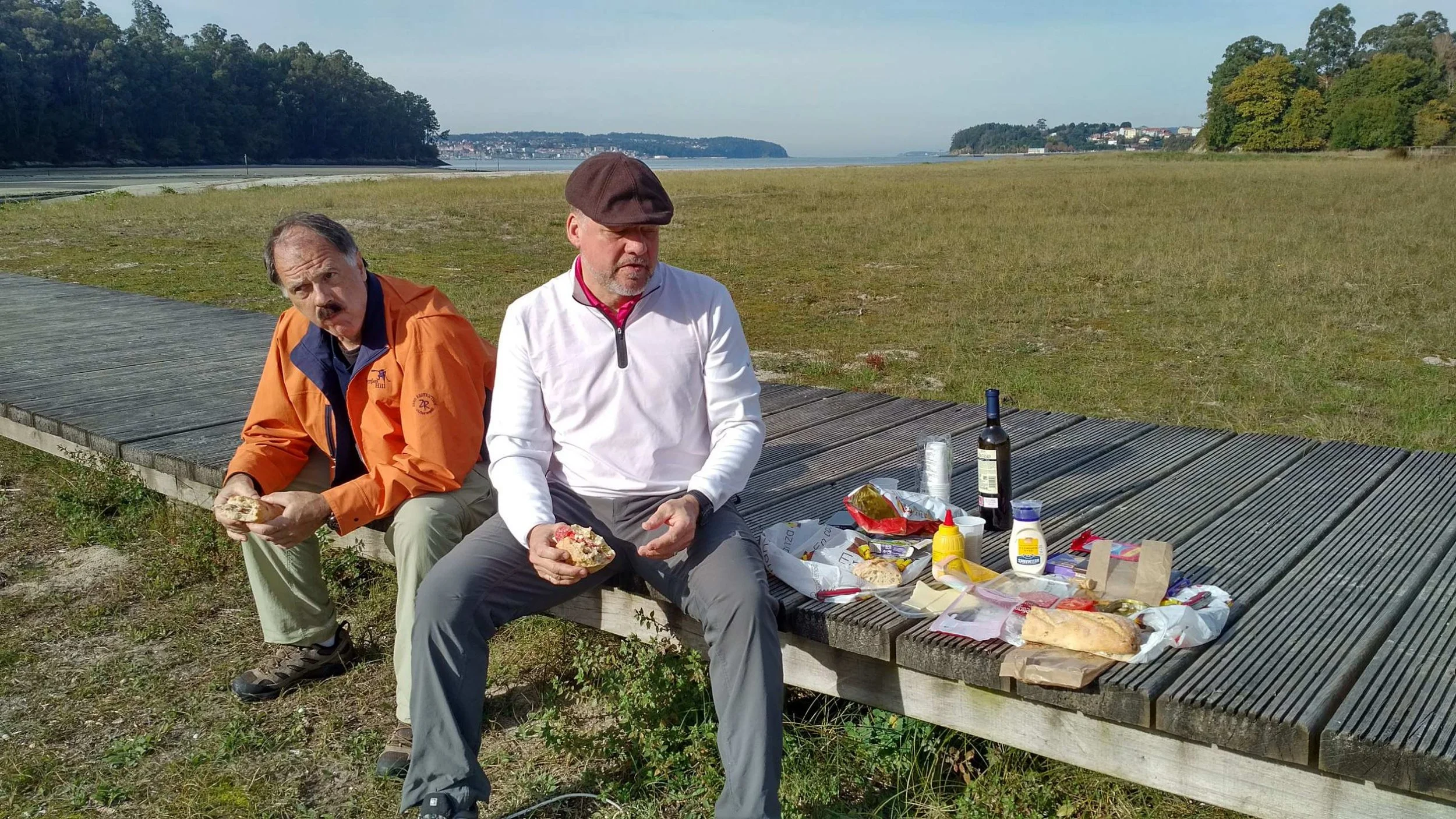 Two men sitting on a wooden bench having a picnic outdoors near a body of water and green hilly landscape, with various food items and condiments on the ground.