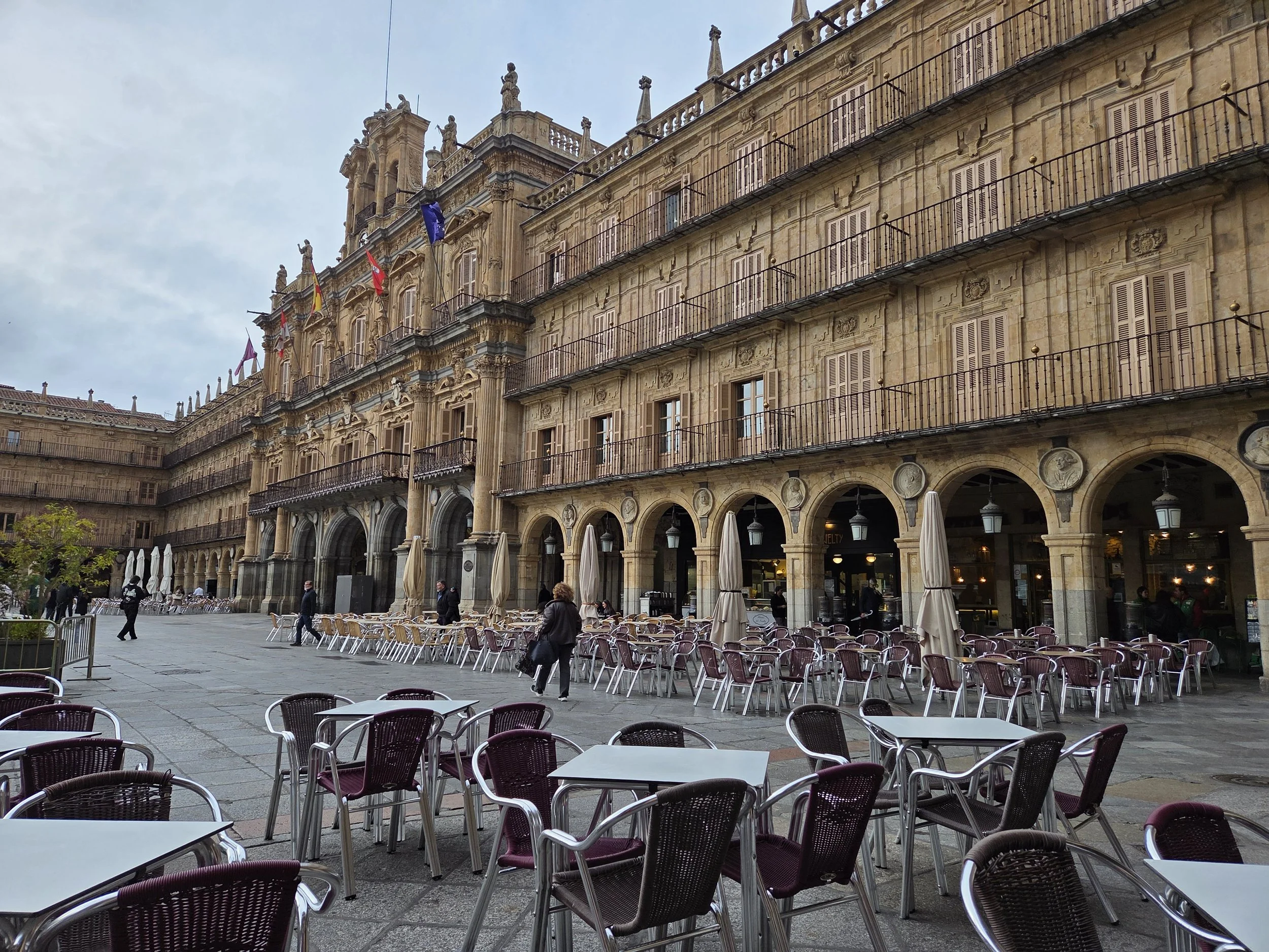 The historic Plaza Mayor in Salamanca showing a building with flags and statues on its façade, overlooking an outdoor cafe with empty tables and chairs.