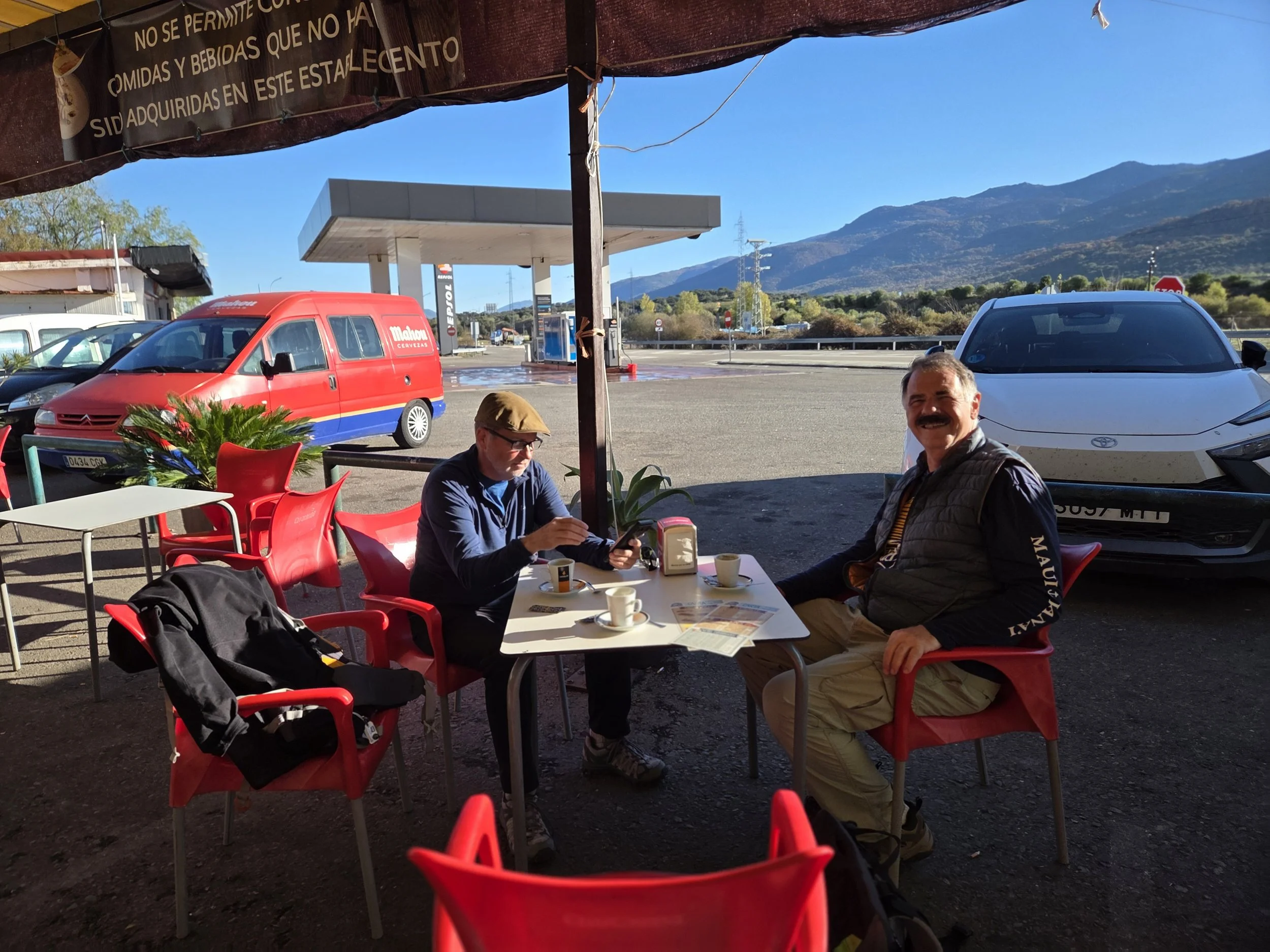 Pilgrims sitting at an outdoor cafe with a mountain in the background, a red van and a white car parked by a gas station, and a cover with Spanish writing overhead on the Via de la Plata.