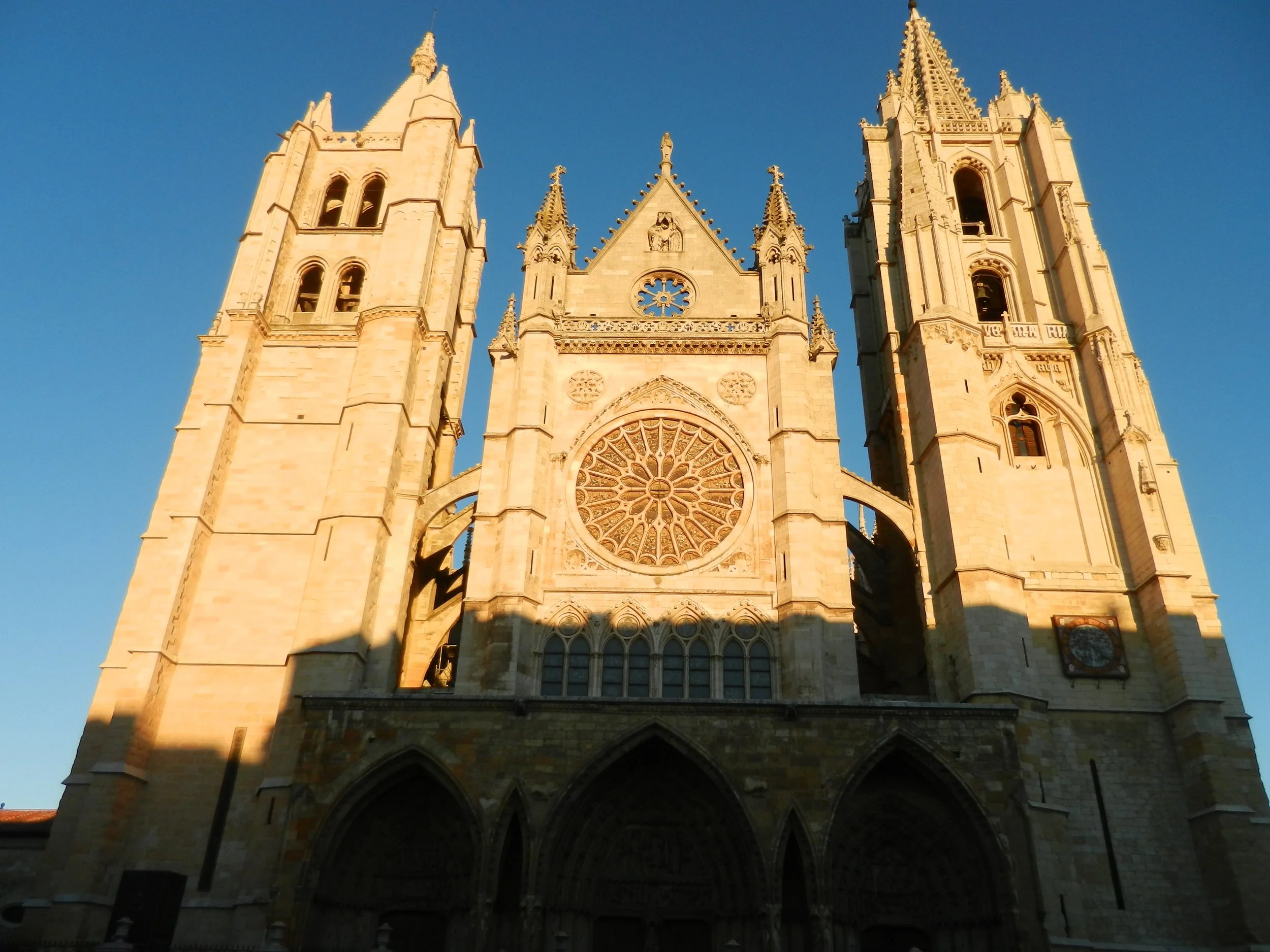 Front view of a Gothic-style cathedral with twin towers, intricate stained glass window, and detailed stone carvings, bathed in golden sunlight against a clear blue sky.