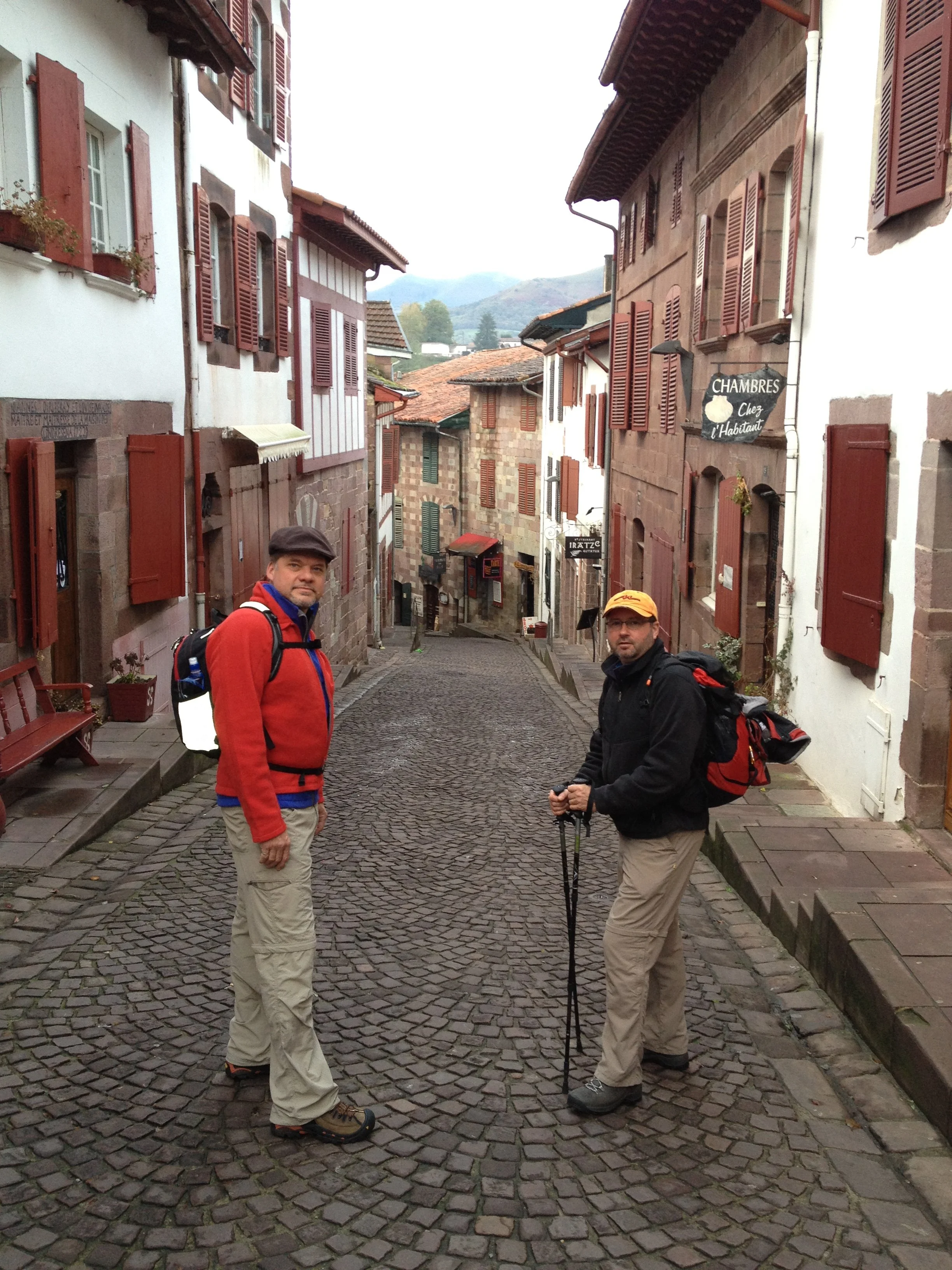 Two men with backpacks standing on a cobblestone street in a European village, surrounded by brick and white buildings with red shutters, and mountains in the background.