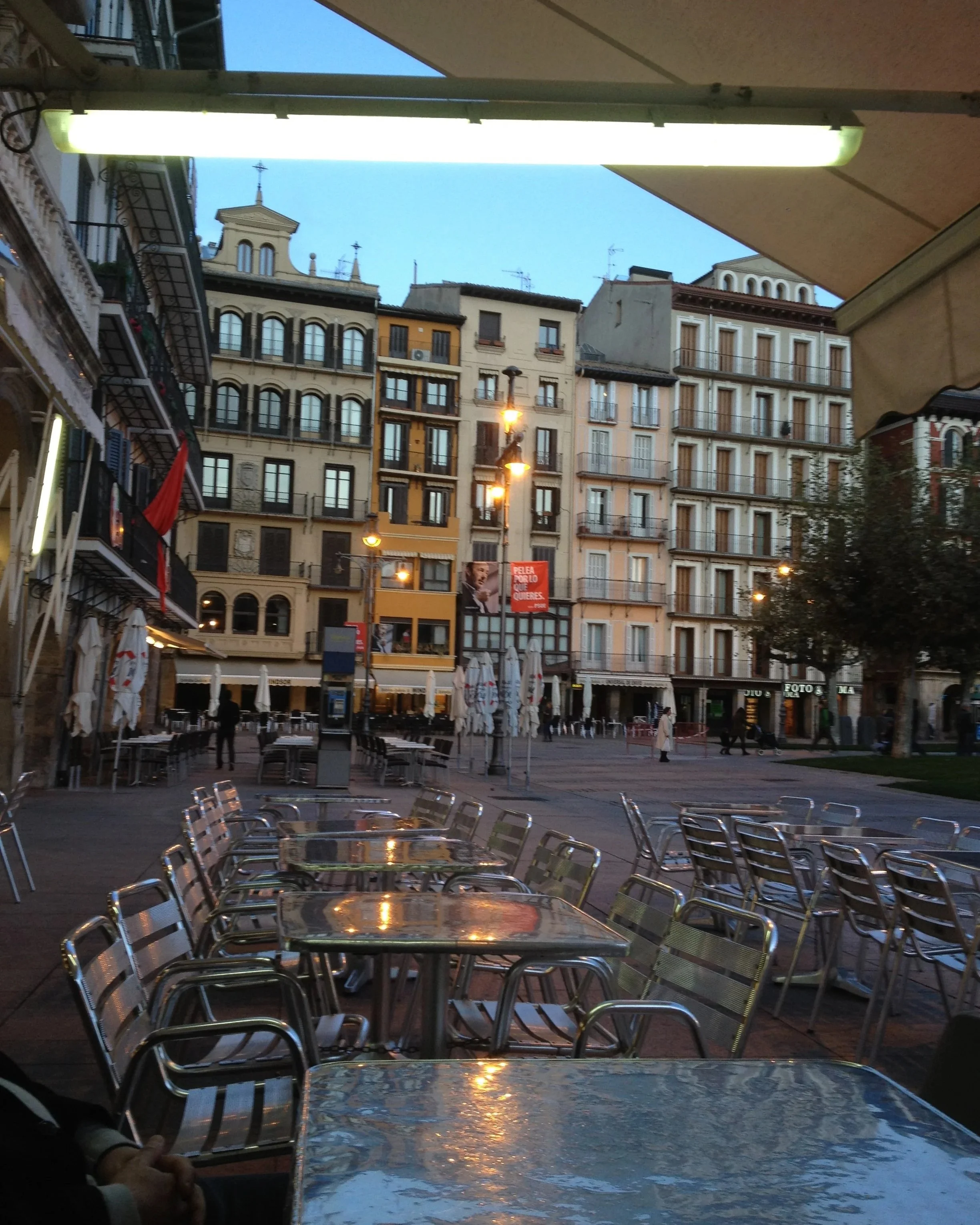 Empty outdoor cafe area with tables and chairs, city buildings in the background, evening sky, and streetlights.