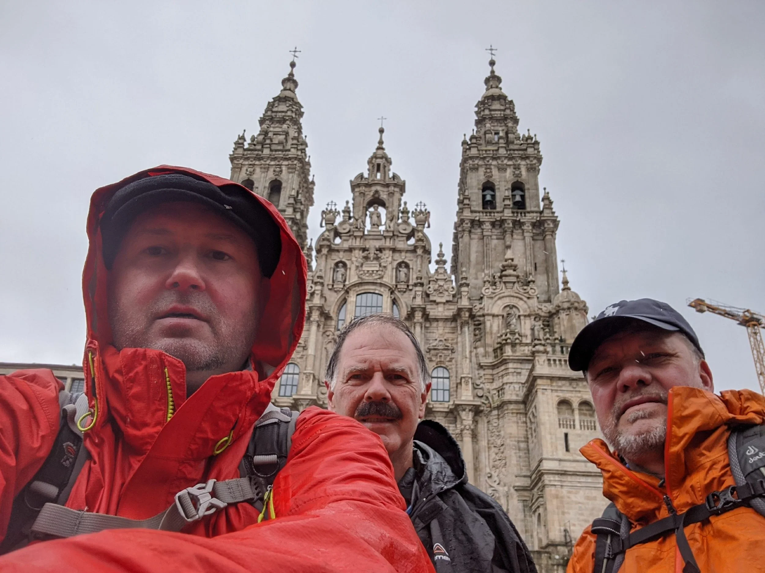 Group of three pilgrims people taking a selfie in front of the cathedral in Santiago de Compostela.   The sky is overcast and the individuals are wearing outdoor gear.