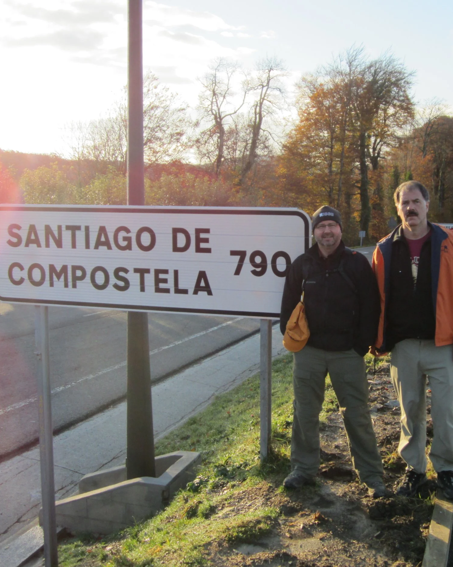 Two men standing next to each other outside near a road, with a road sign indicating Santiago de Compostela at 790 kilometers. The men are dressed in outdoor clothing, and the background features trees with fall foliage.