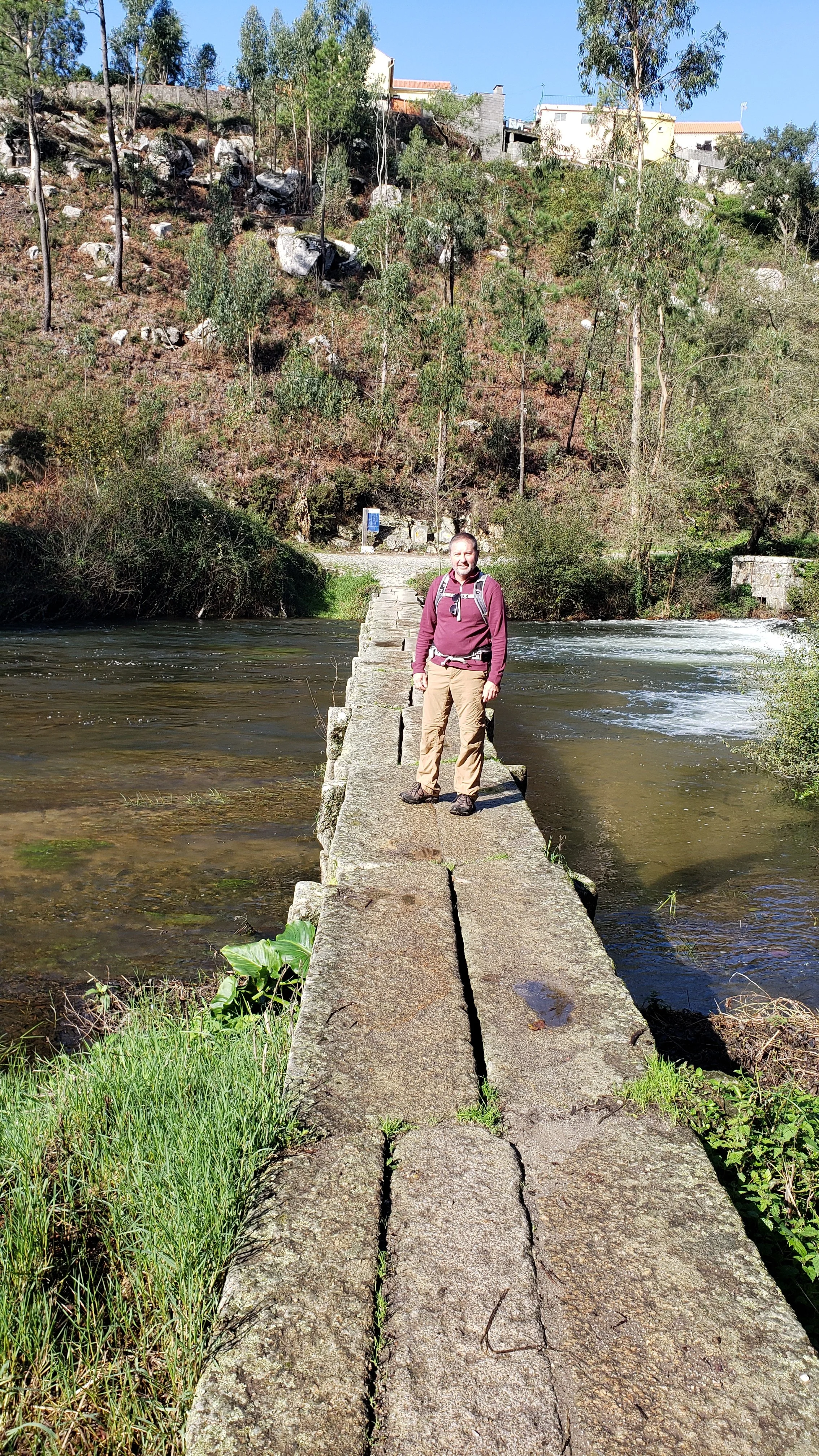A man standing on a narrow stone pedestrian bridge over a river, with trees and houses on a hillside in the background.