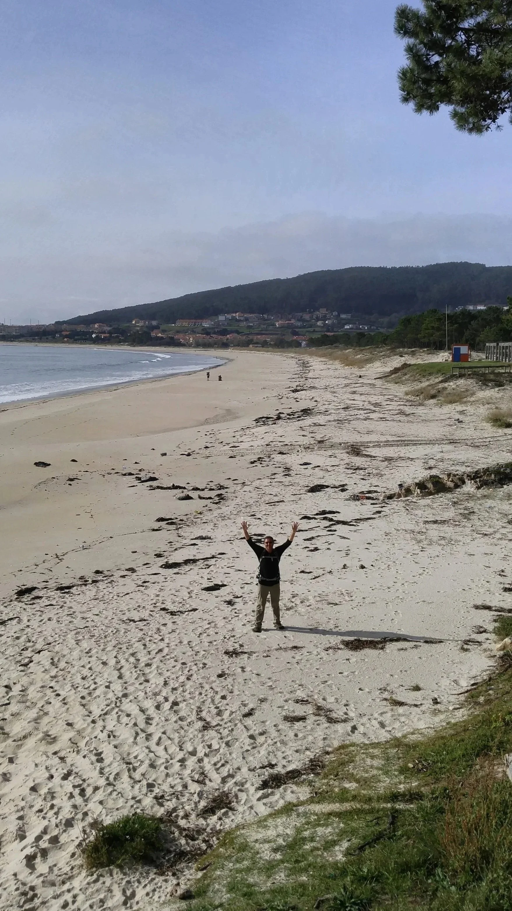 Person standing on the Praia de Langosteira, near Finisterre, with arms raised, ocean waves to the left, and a sandy shoreline to the right, with green vegetation in the foreground and a distant hillside in the background.