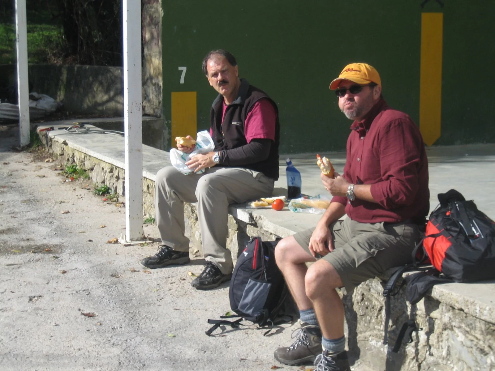 Two men sitting on a concrete bench outdoors, eating sandwiches, enjoying a Pilgrim lunch on the Camino. They have backpacks placed on the ground beside them.