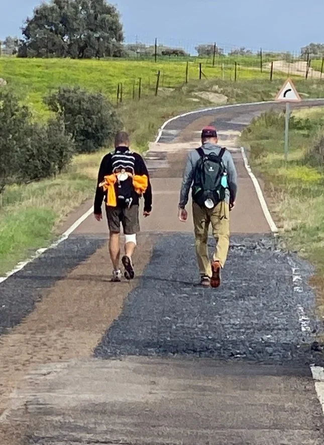 Two hikers walking along a rural asphalt road with cracks and patches, surrounded by green fields and a few trees, with a warning sign ahead indicating a curve.