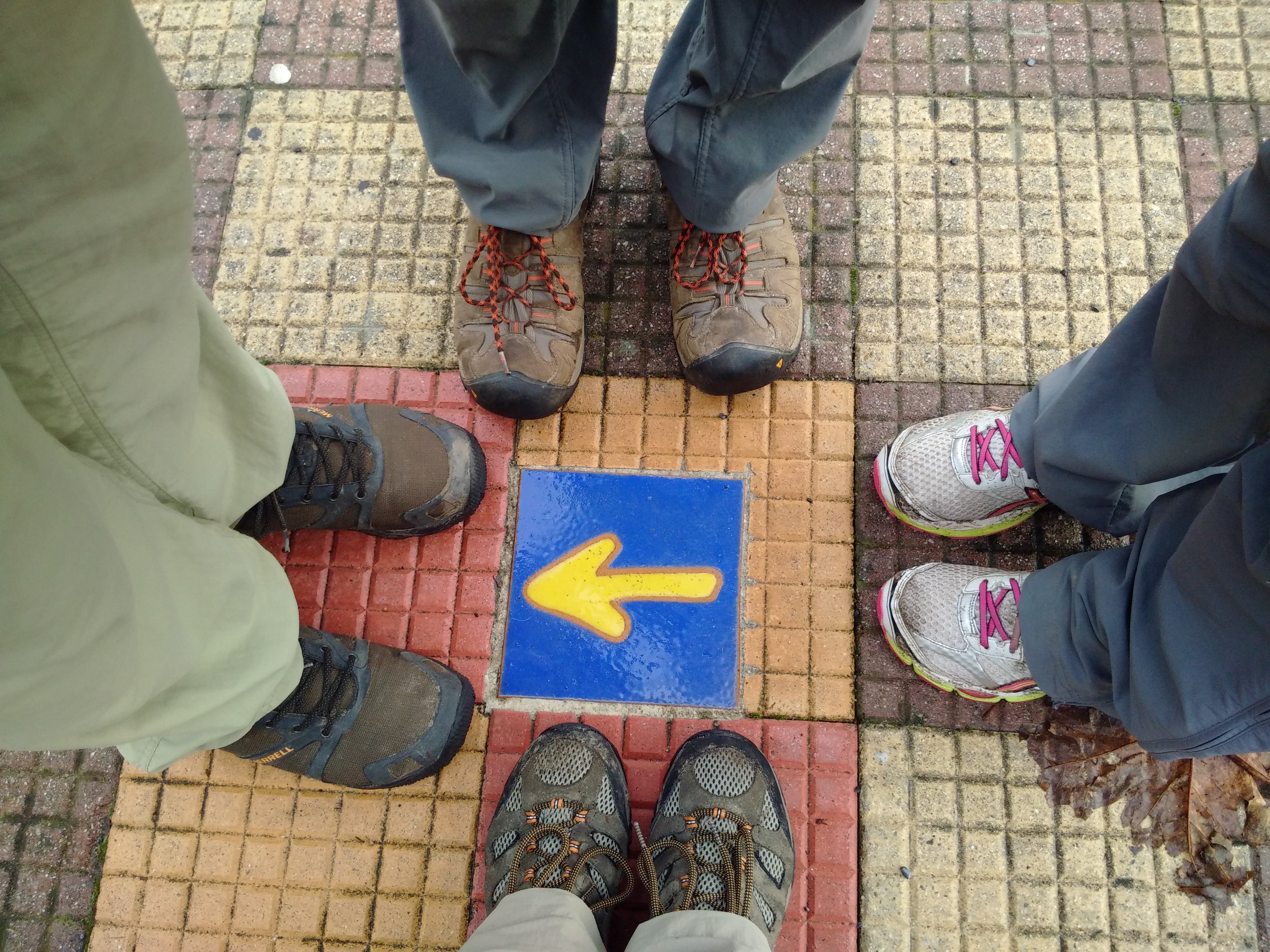 View from above of four pairs of hiking shoes on a brick-painted sidewalk, with a blue and yellow Camino directional arrow painted on the pavement.
