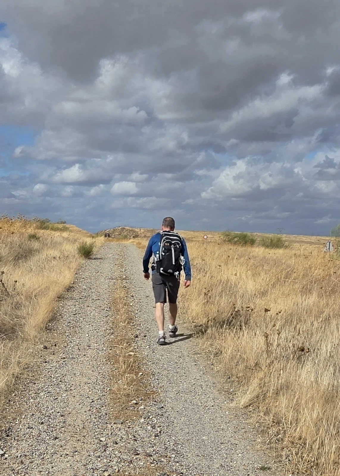 A Camino pilgrim hiking on a dirt trail through a dry, grassy landscape under a cloudy sky on the Via de la Plata.