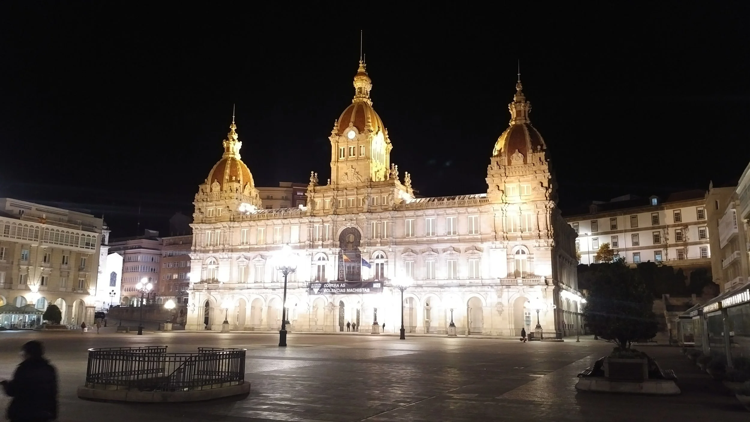 Night view of a historic building with three domed towers, illuminated and surrounded by city buildings and empty town square.