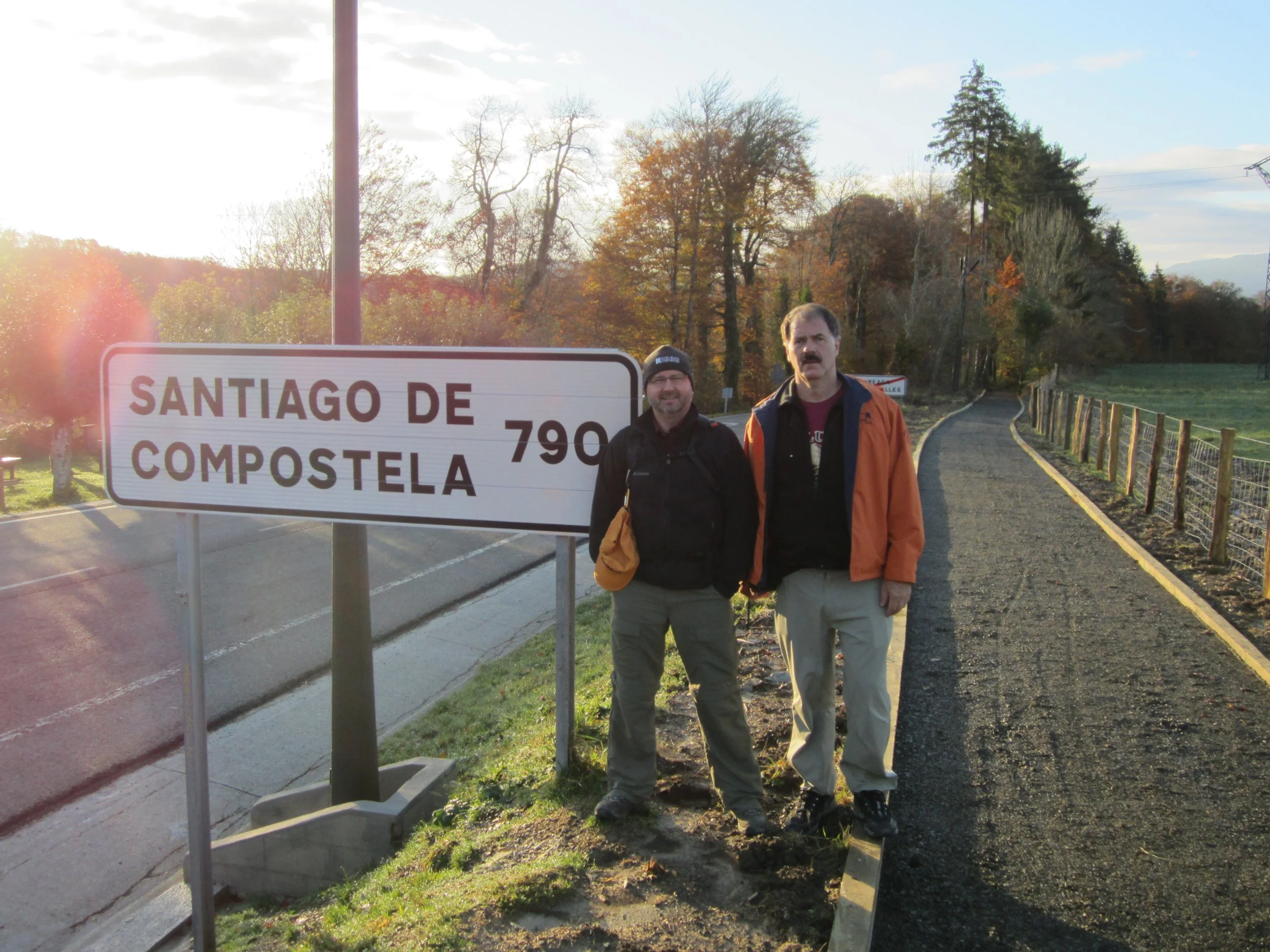 Two men standing beside a road sign that reads "Santiago de Compostela" with autumn trees in the background and a partially cloudy sky.