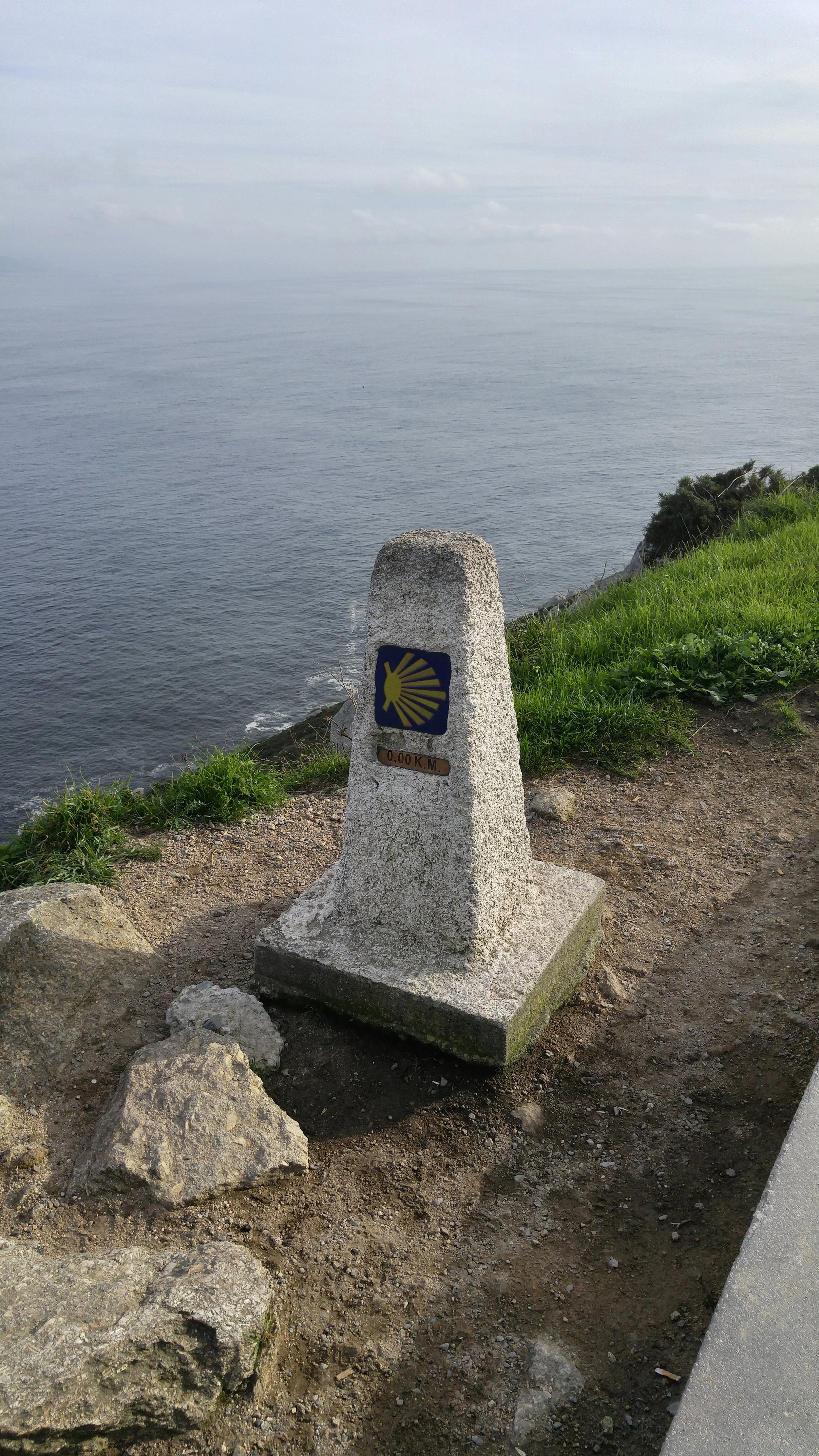 A stone marker along a coastal trail with a yellow scallop shell symbol and a sign indicating zero kilometers, near the ocean with a grassy area and rocks.