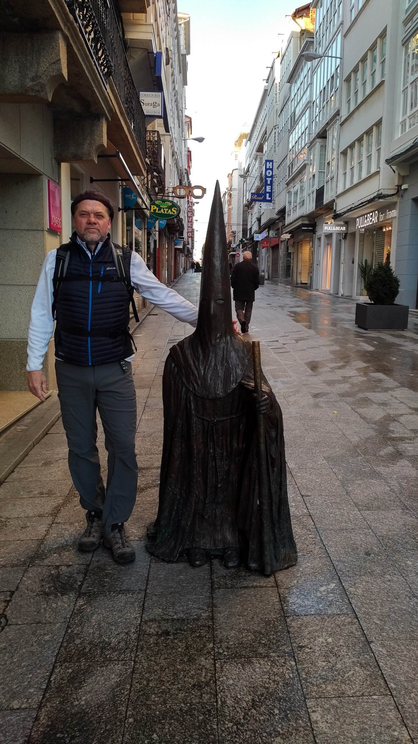 A man standing next to a wooden sculpture of a mythical creature with a long, pointed nose, in a city street with shops and buildings.