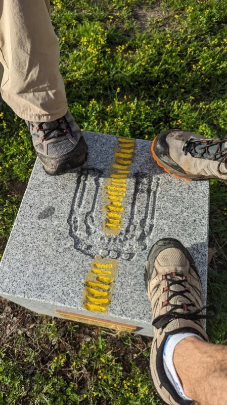 Two people in hiking shoes standing on a stone marker with yellow and black markings in a grassy area with small yellow flowers.