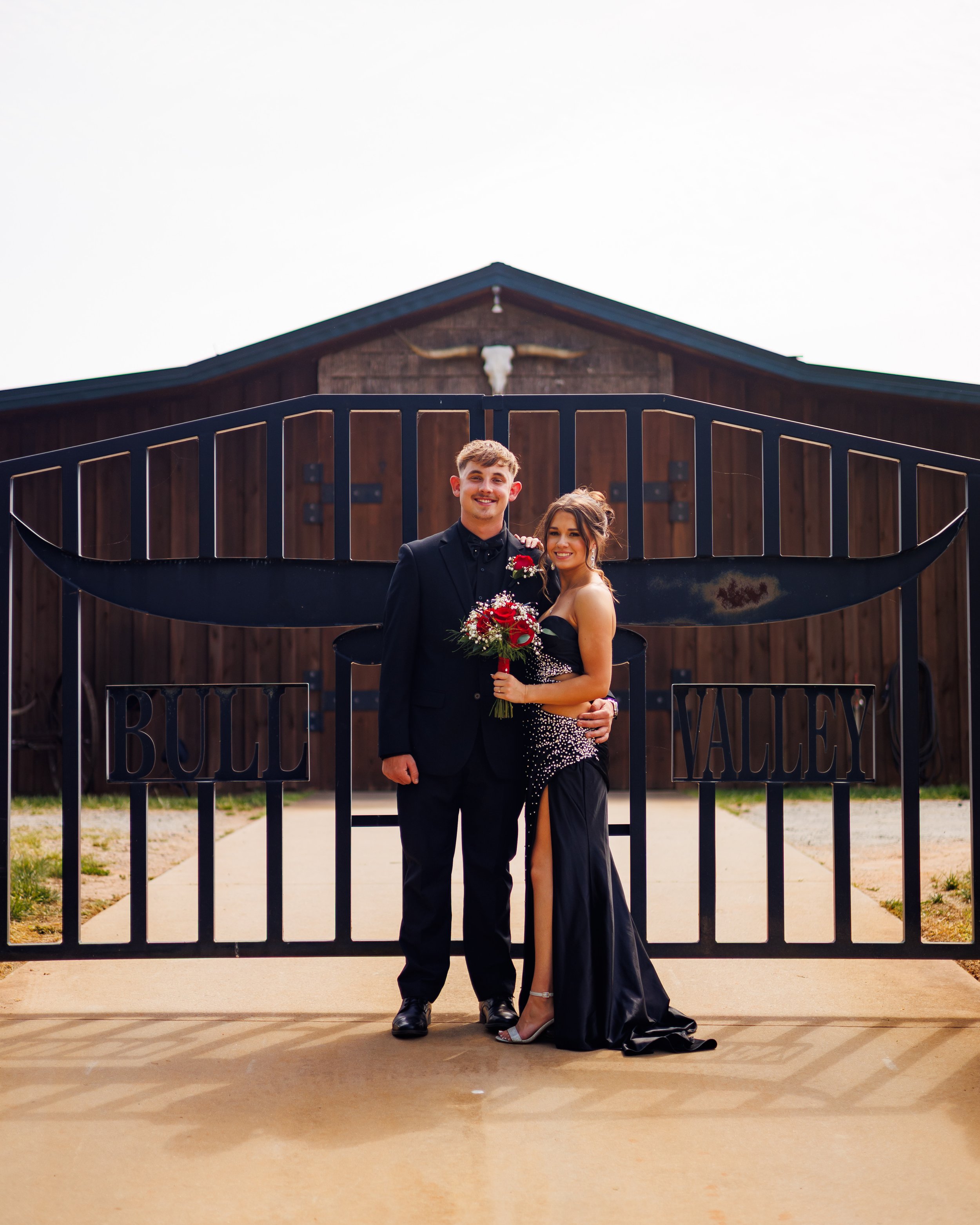 A young man and woman in formal attire standing together behind a gate with a barn in the background. The man is wearing a black tuxedo and the woman is in a black evening gown holding a bouquet of red and white flowers.