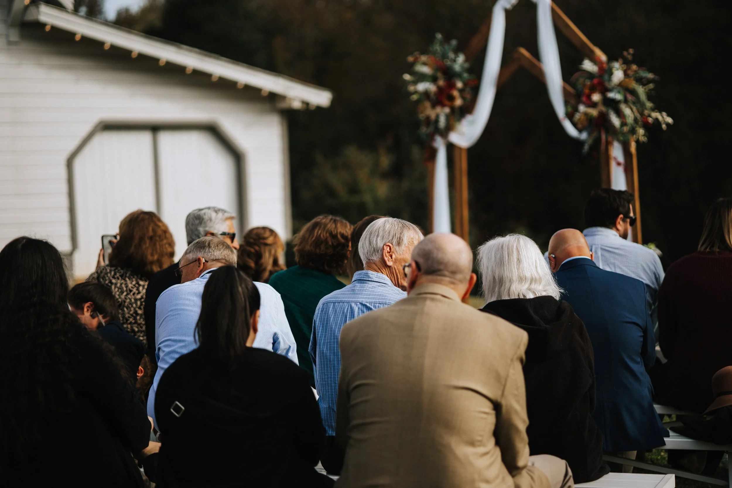 A crowd of people gathered outdoors near a small white building with a gabled roof, decorated with string lights. In the background, there is an arch adorned with flowers and white draping, likely for a special event or ceremony.