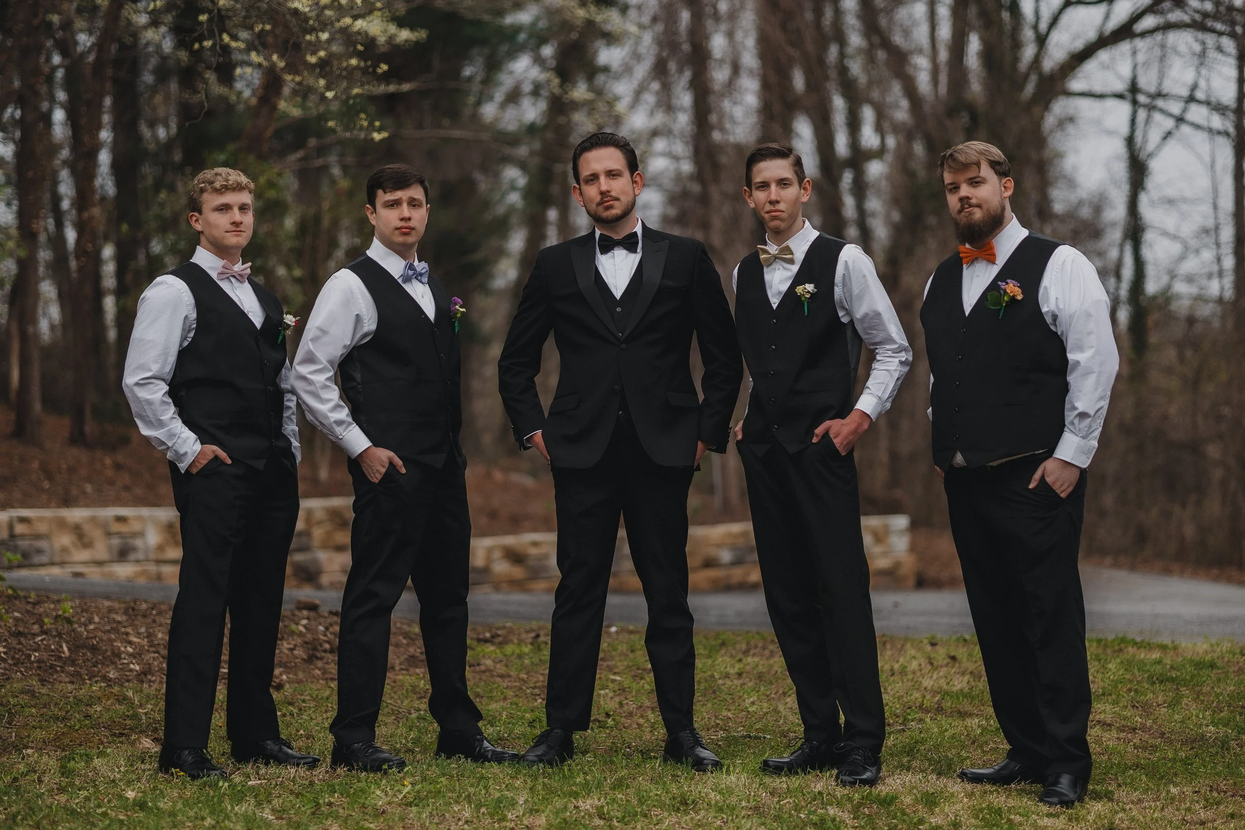 Five men in formal attire, standing outdoors among trees, posing confidently for a photo. They are wearing black vests, white shirts, and bow ties, with the man in the center dressed in a tuxedo.