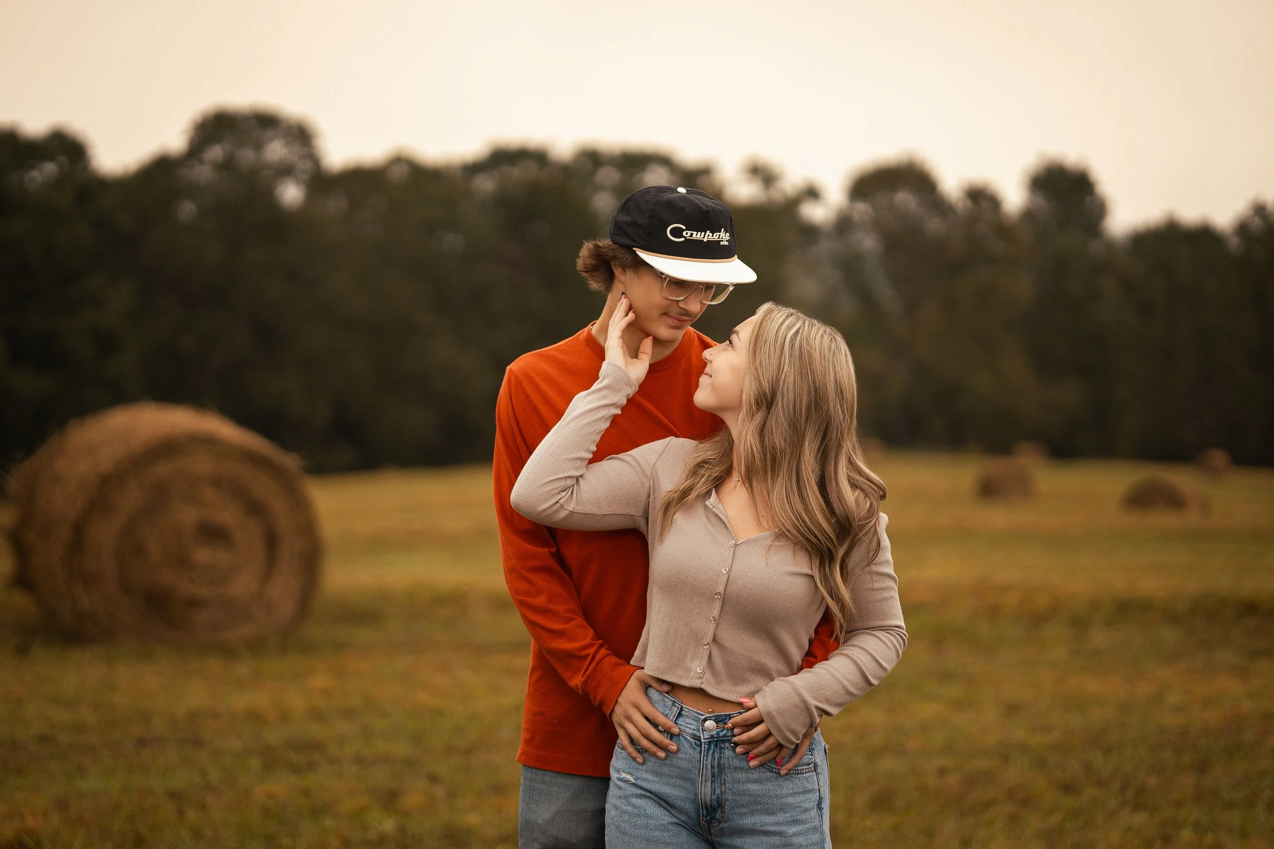 A couple stands close together outdoors on a grassy field with hay bales, embracing and looking into each other's eyes during sunset or late afternoon.