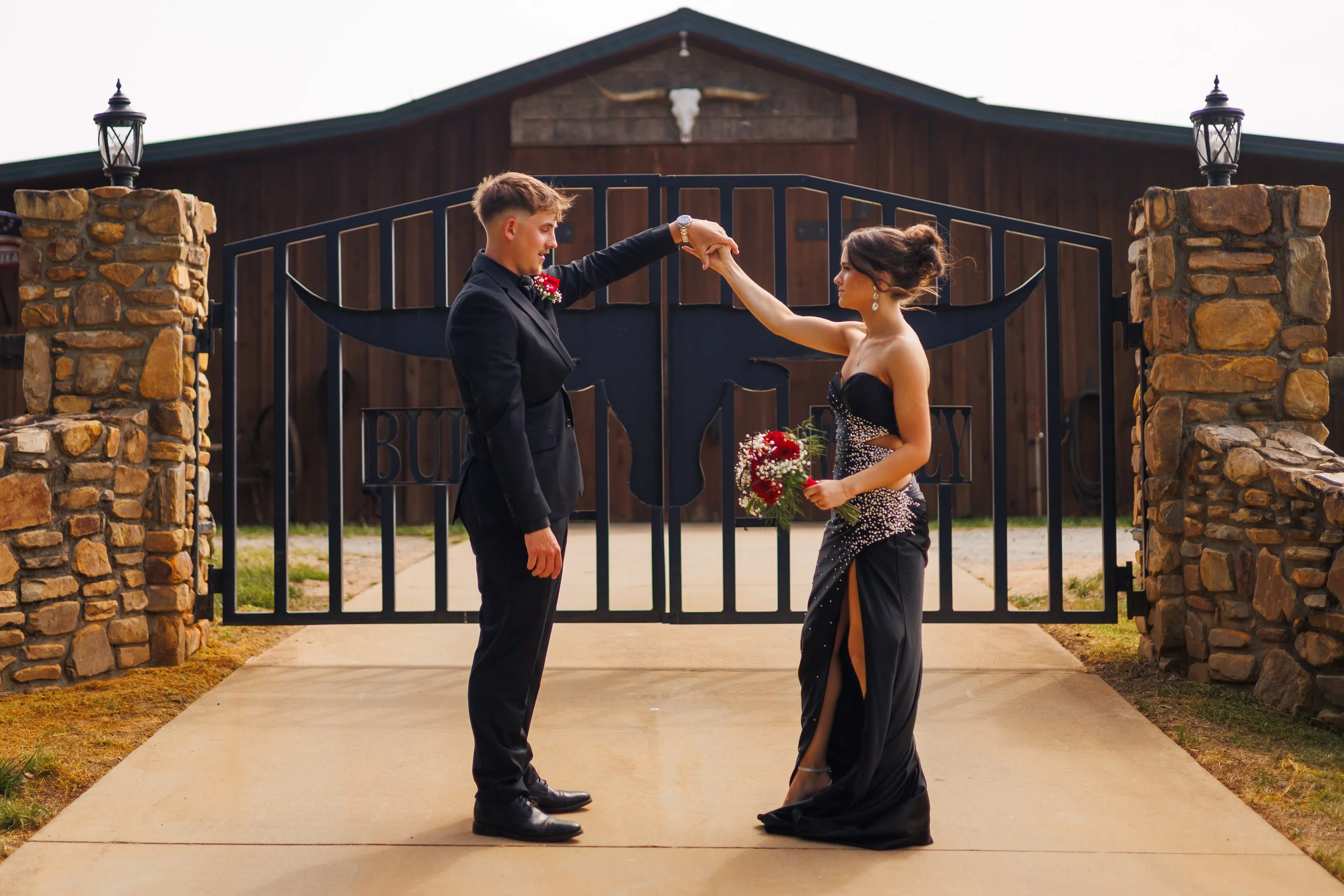 A man in a black suit and a woman in a black dress are standing outside near a gate with stone pillars, holding hands and looking at each other, with a barn in the background.