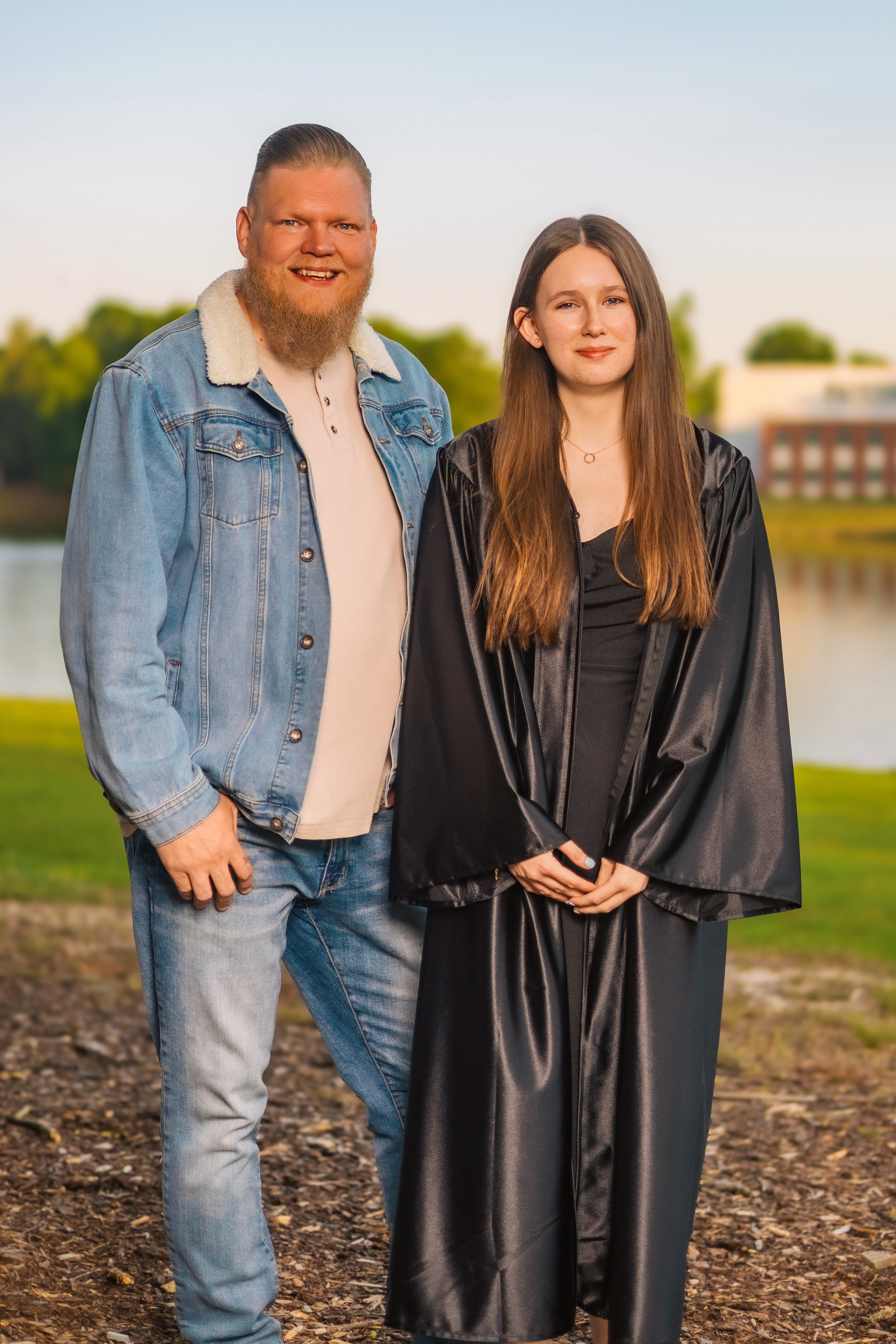 A young woman in a black graduation gown and a man in casual denim clothing standing outdoors near a body of water with trees and buildings in the background.