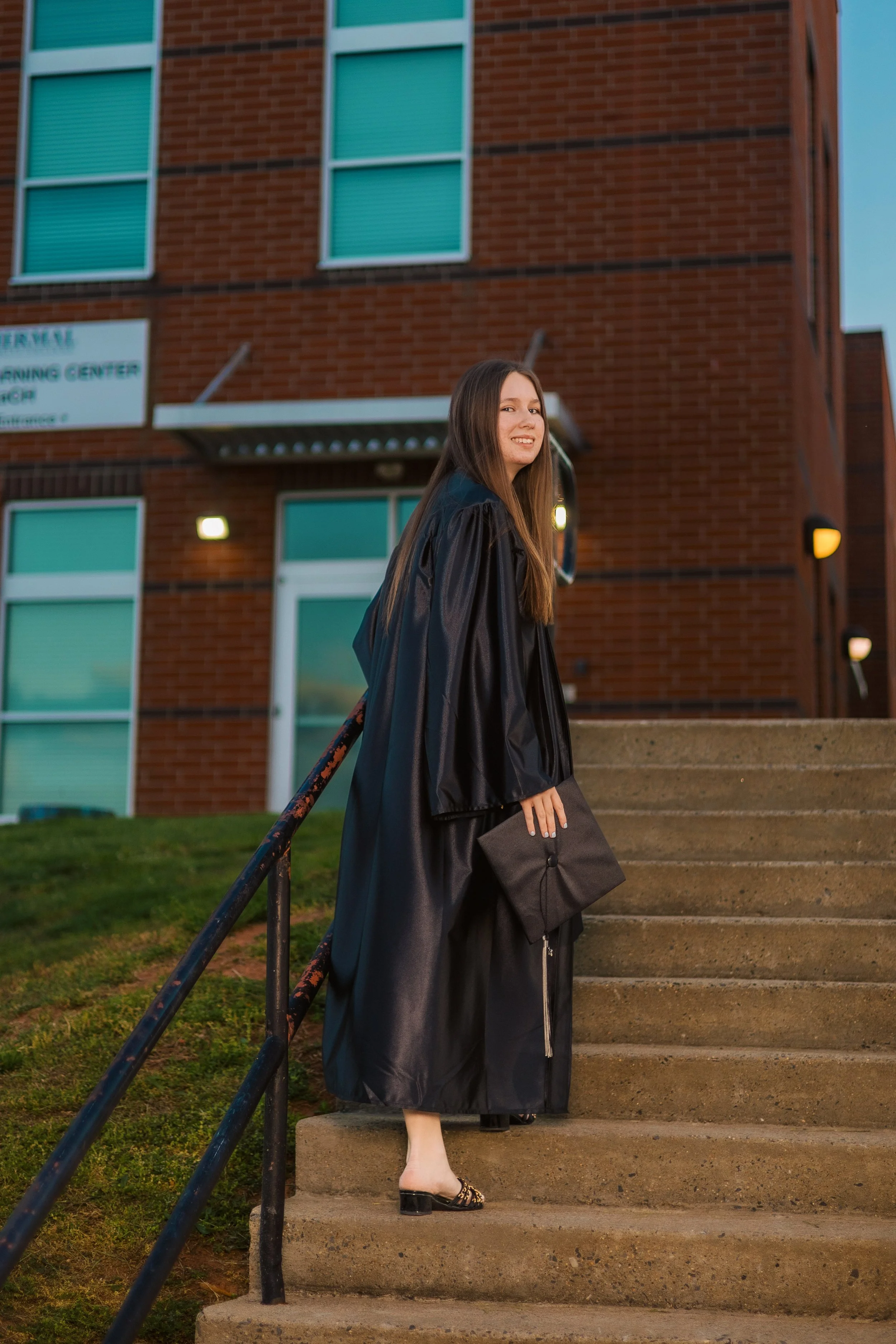 A young woman in a black graduation gown standing on concrete steps outside a brick building, smiling at the camera. The building has windows with teal window coverings and a sign partially visible on the wall.