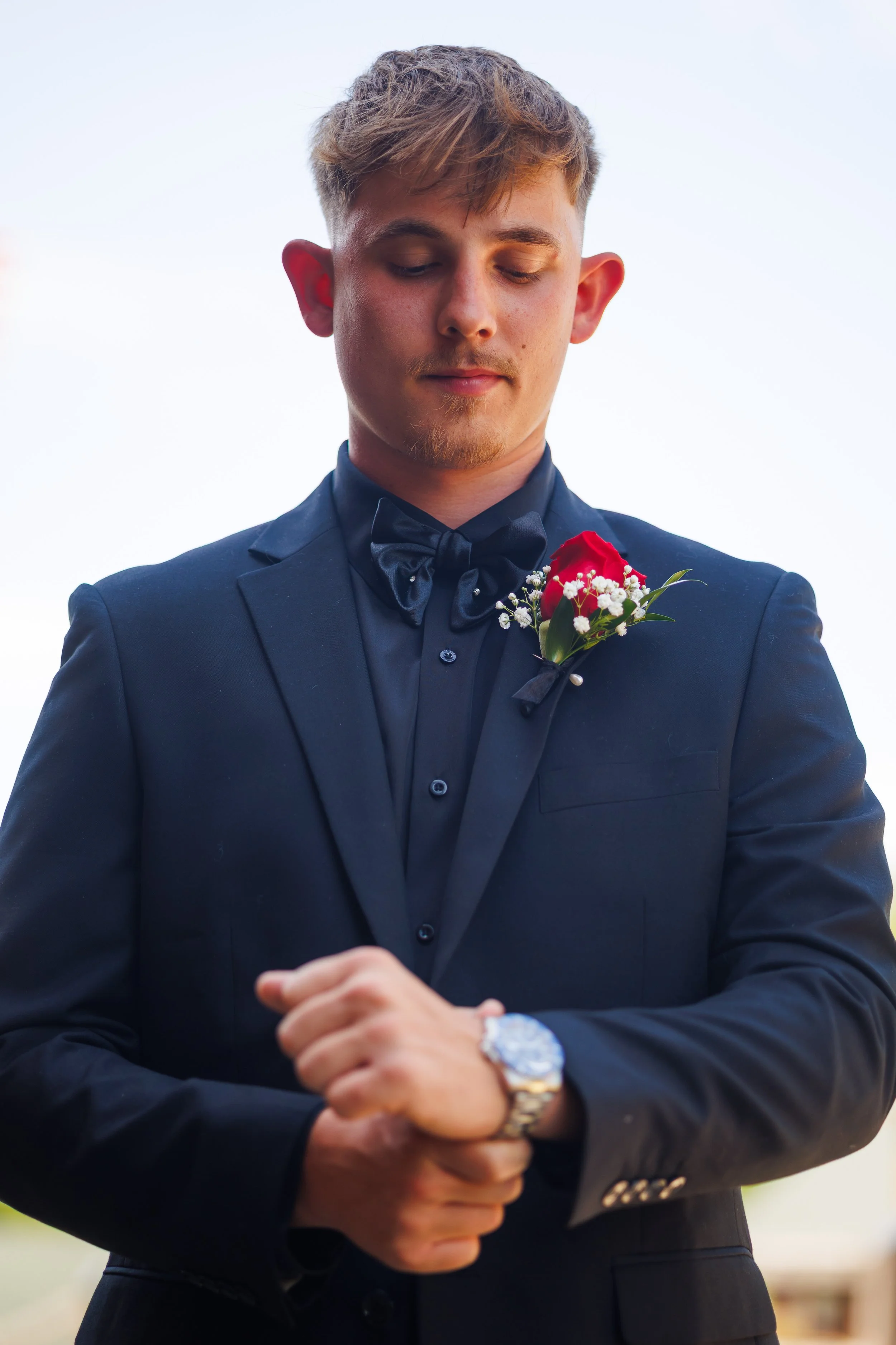 A young man dressed in a dark tuxedo with a black bow tie and a floral boutonniere, standing outdoors with his eyes closed and hands clasped, possibly at a formal event or wedding.