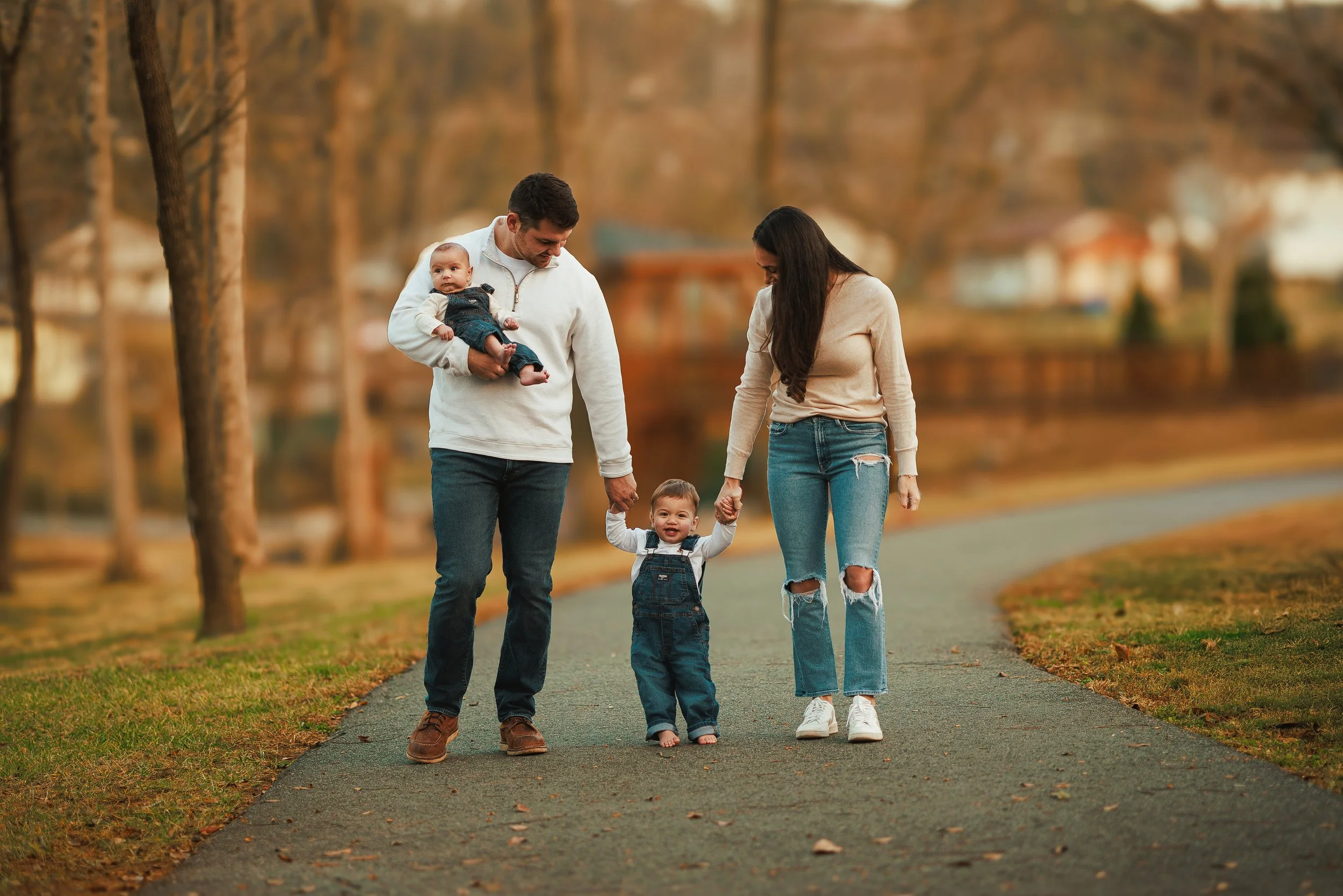 A family of five walking outdoors in a park with autumn trees. The father is holding a baby, and a young child is holding hands with the mother, all smiling and enjoying a fall walk.