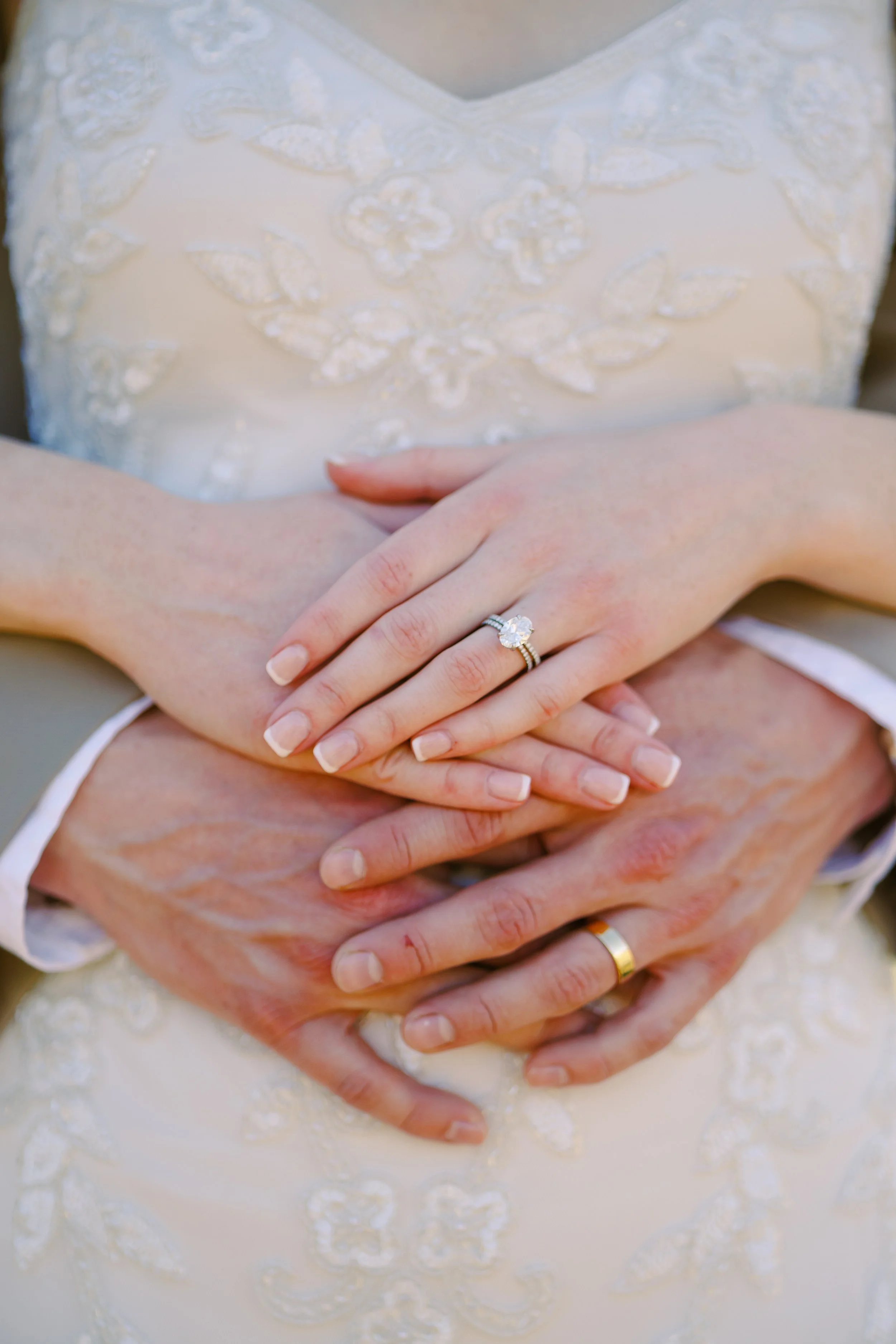 Close-up of a bride and groom holding hands during their wedding, with the bride wearing a white dress with lace embroidery and the groom wearing a tan suit.
