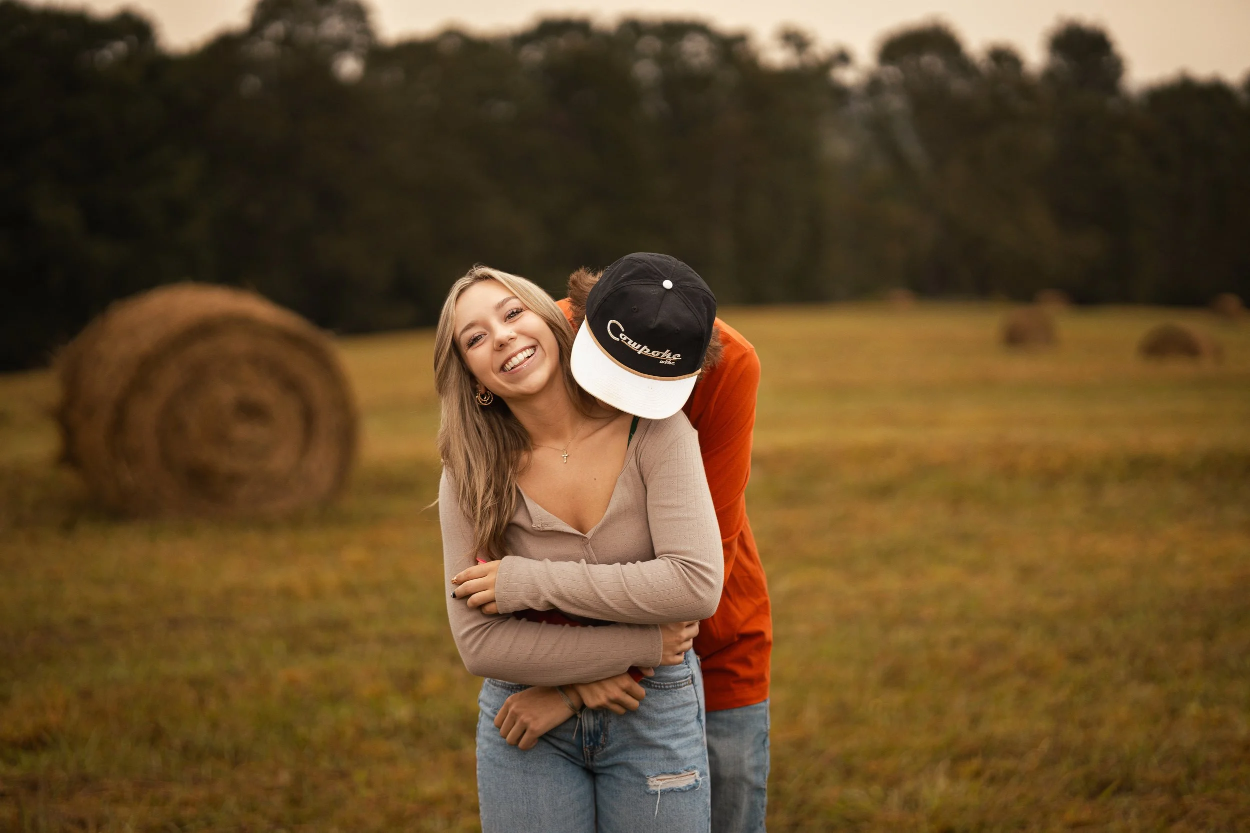 A woman stands in a field with hay bales, smiling, while a person behind her leans over her shoulder, wearing a black and white cap. The scene is outdoors during sunset with trees in the background.