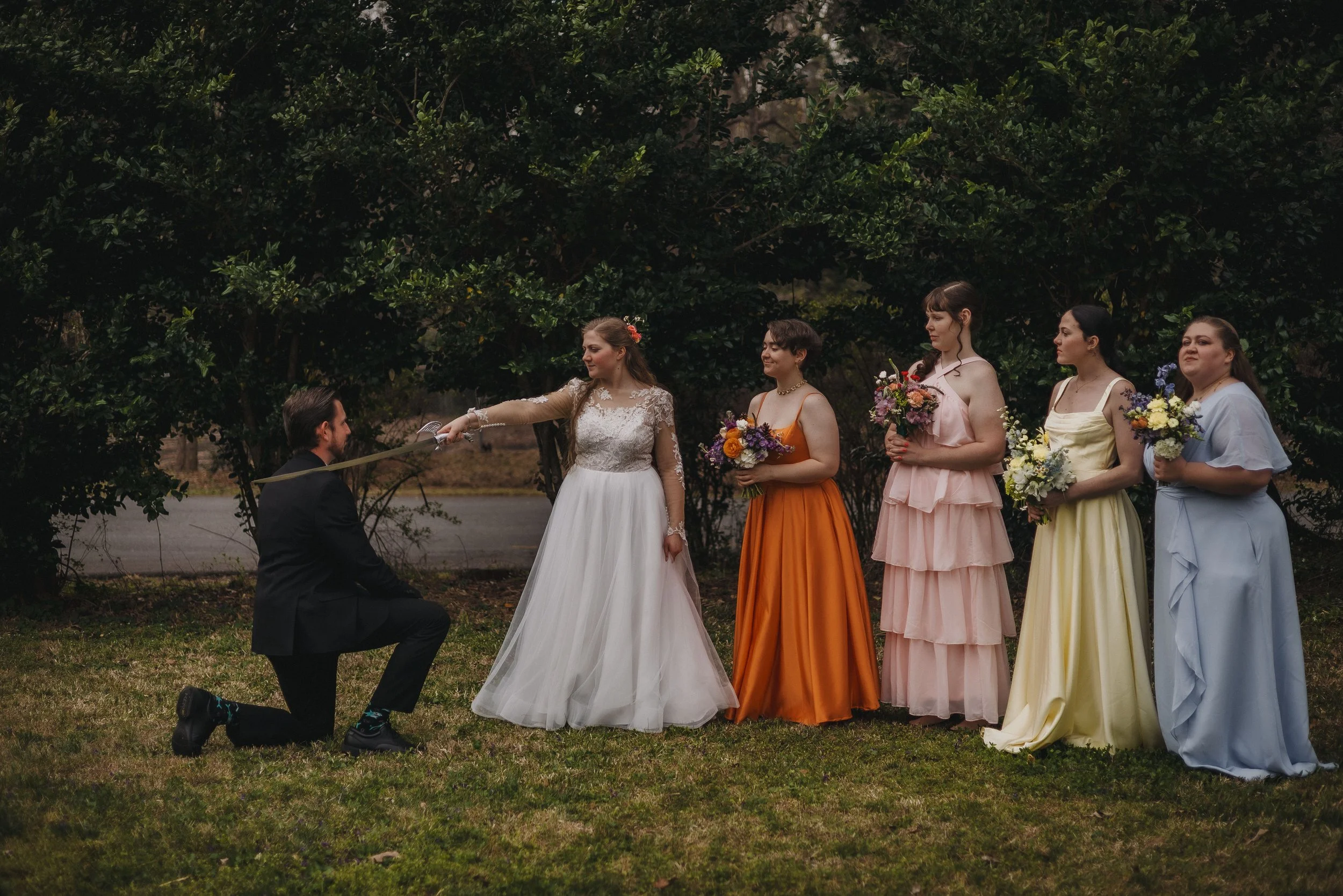 A wedding ceremony outdoors with a bride, groom, and four bridesmaids holding bouquets, standing on grass with trees in the background.