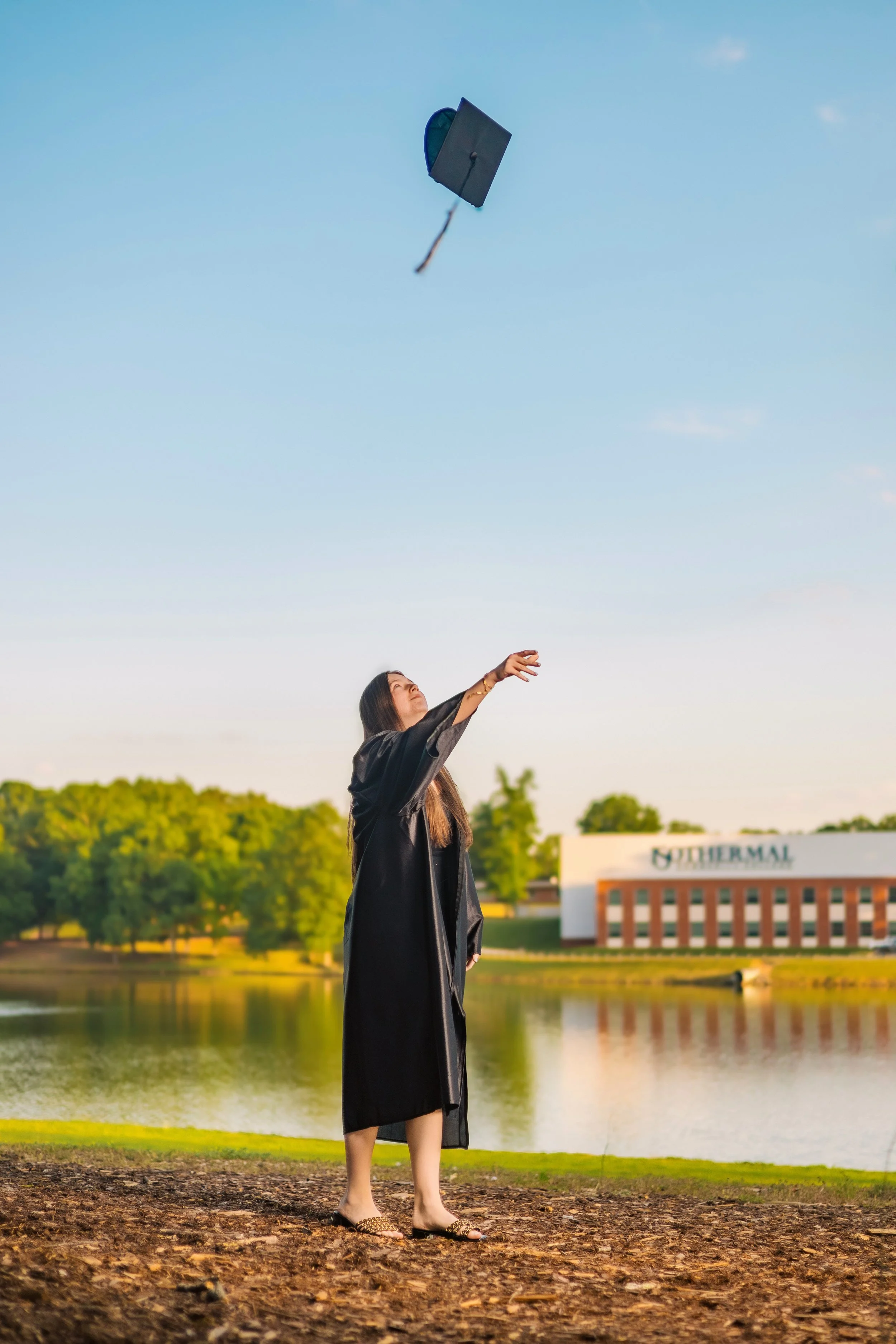 A woman in a black graduation gown and cap, standing outdoors near a body of water, throwing her cap into the sky during her graduation celebration in the evening.