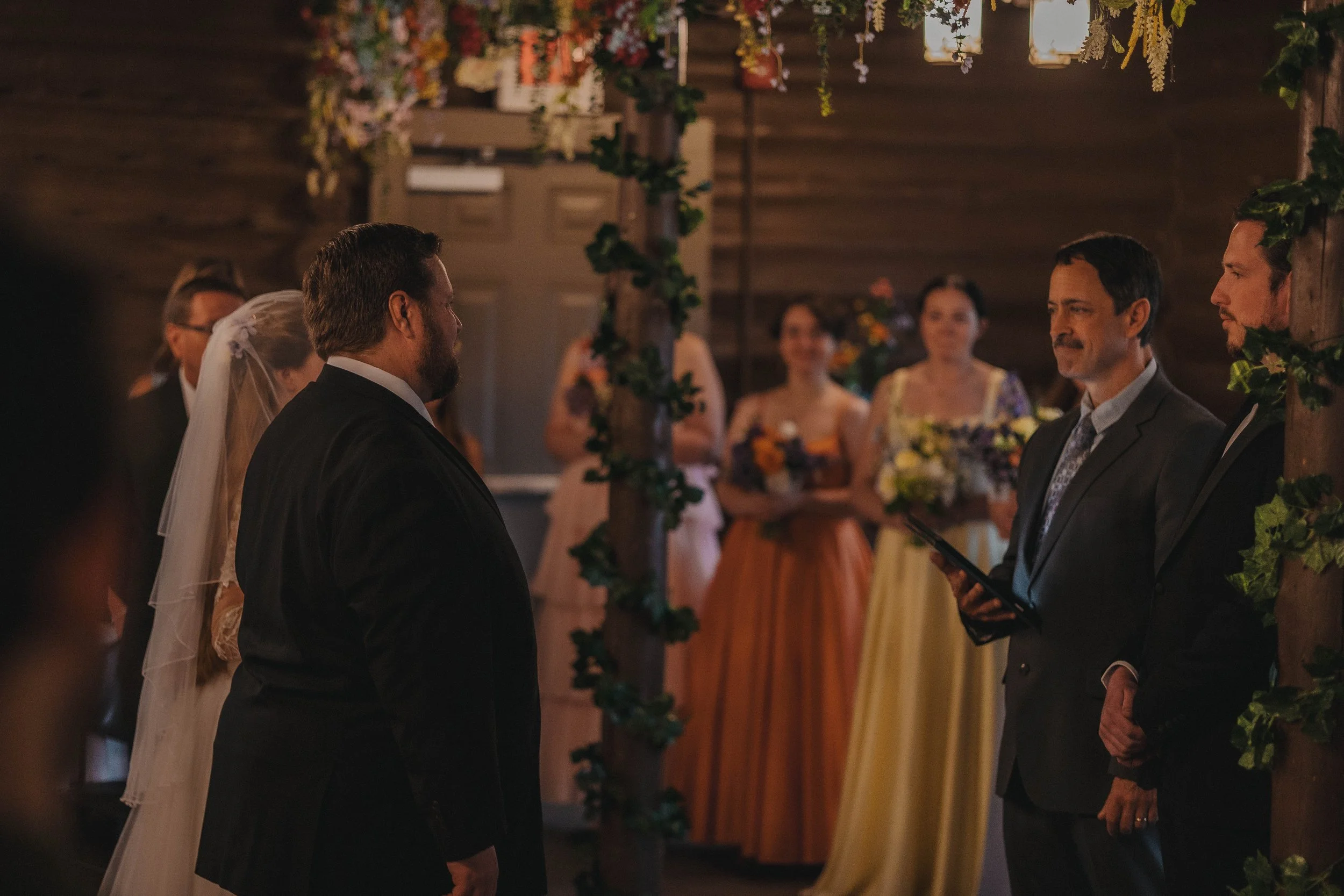 A wedding ceremony with the bride and groom facing each other under an arch decorated with flowers and greenery, with guests witnessing the occasion in a warmly lit wooden venue.
