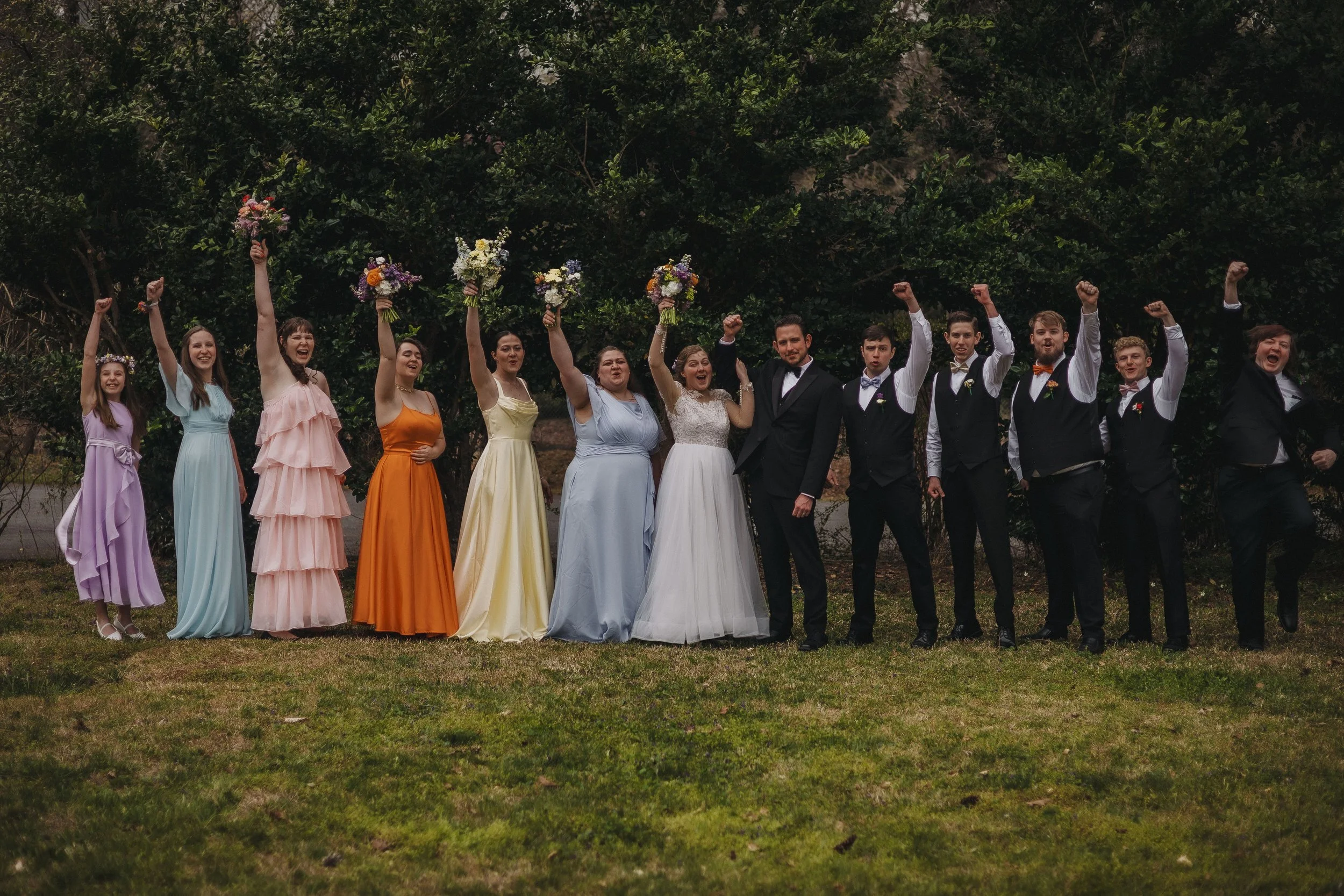 A wedding party standing outdoors on grass, holding bouquets and raising their fists in celebration. The group includes women in colorful dresses and men in formal suits, with lush green trees in the background.