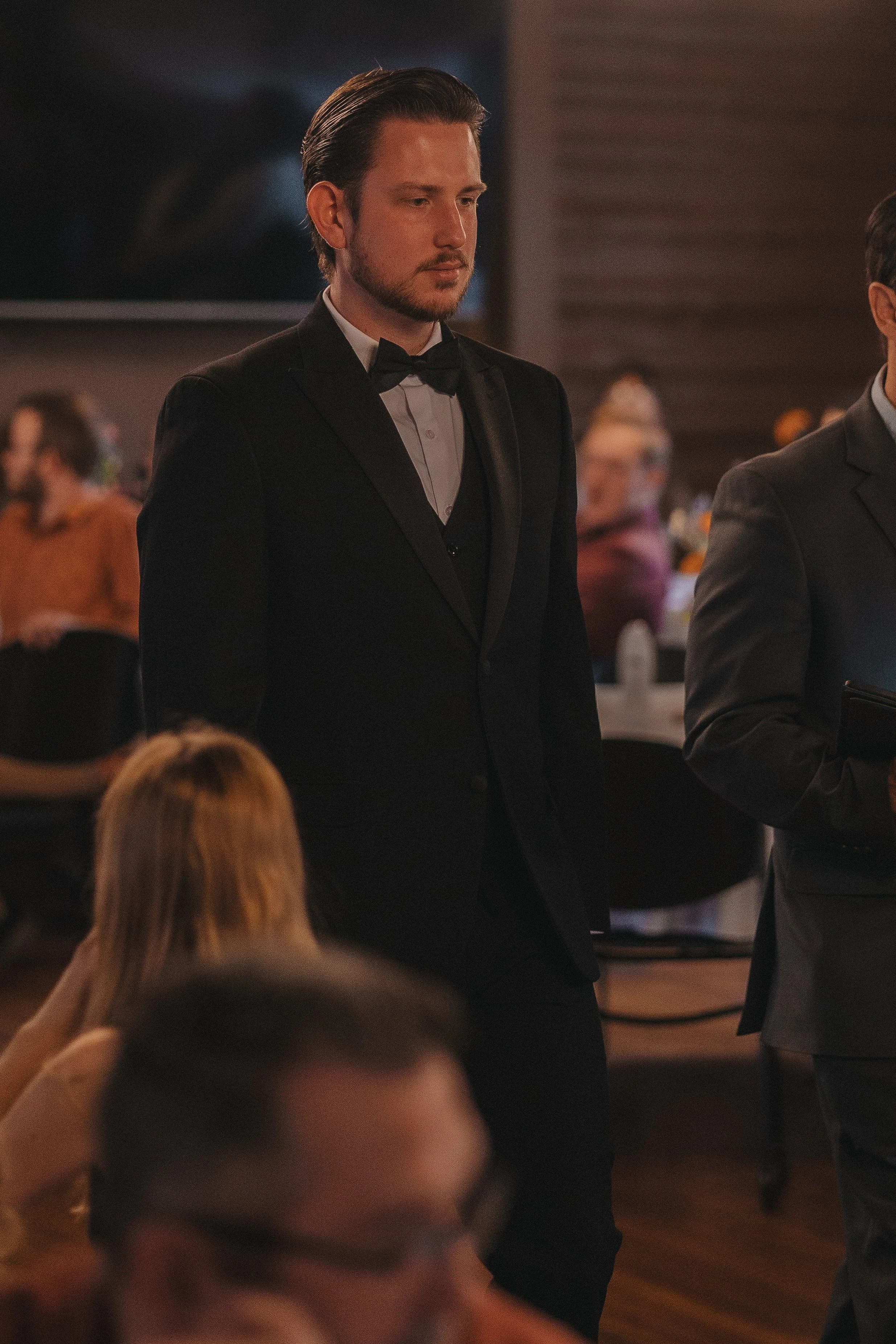 Man in formal black tuxedo with bow tie standing at a formal event or gathering, with other people in the background.