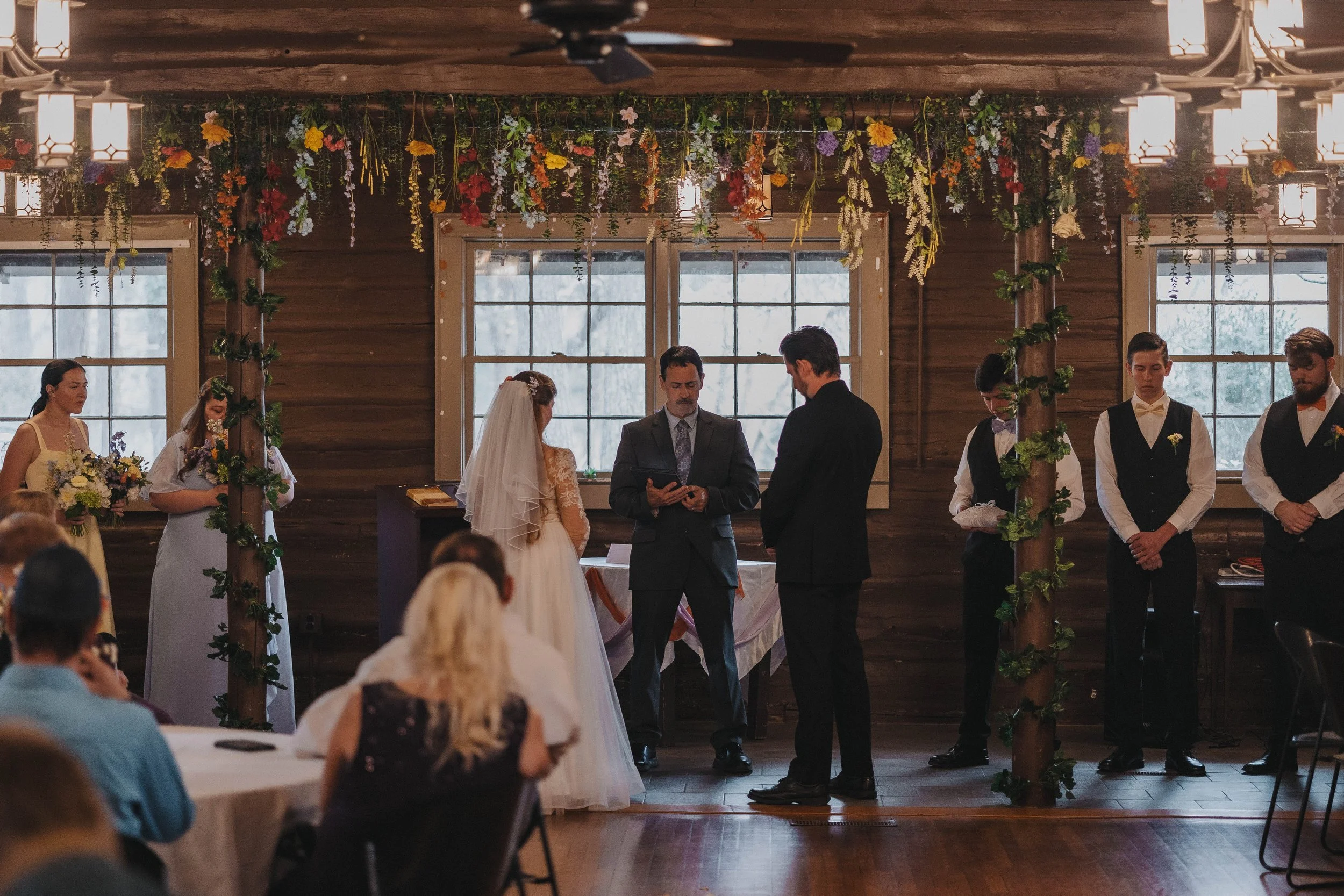 A wedding ceremony taking place inside a rustic wooden venue with large windows, decorated with hanging flowers and greenery. The bride and groom are standing at the altar with a person officiating, while bridesmaids and groomsmen stand nearby. Guest