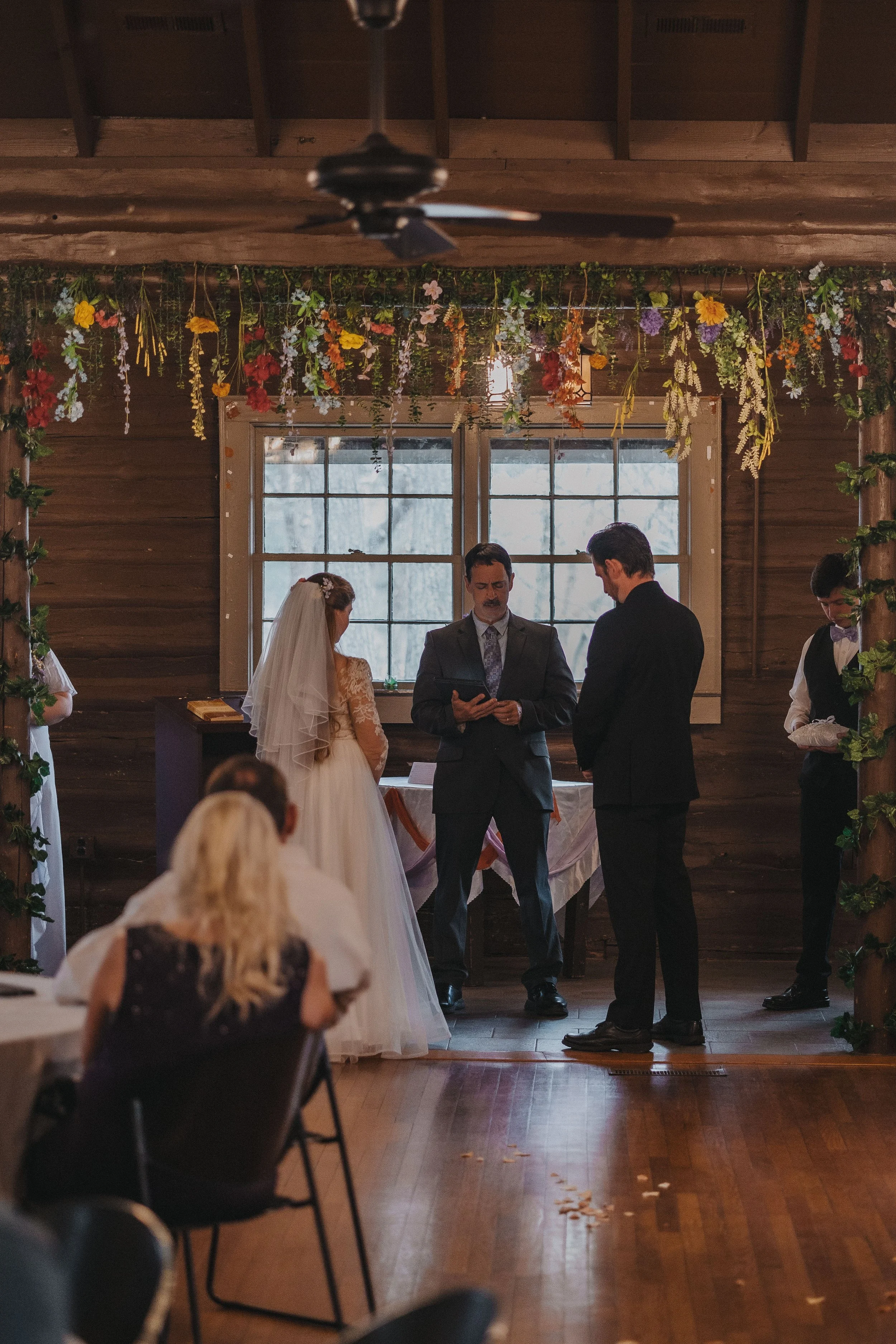 A wedding ceremony inside a rustic wooden venue with floral decorations hanging from the ceiling, a bride and groom standing with an officiant, and guests seated watching.
