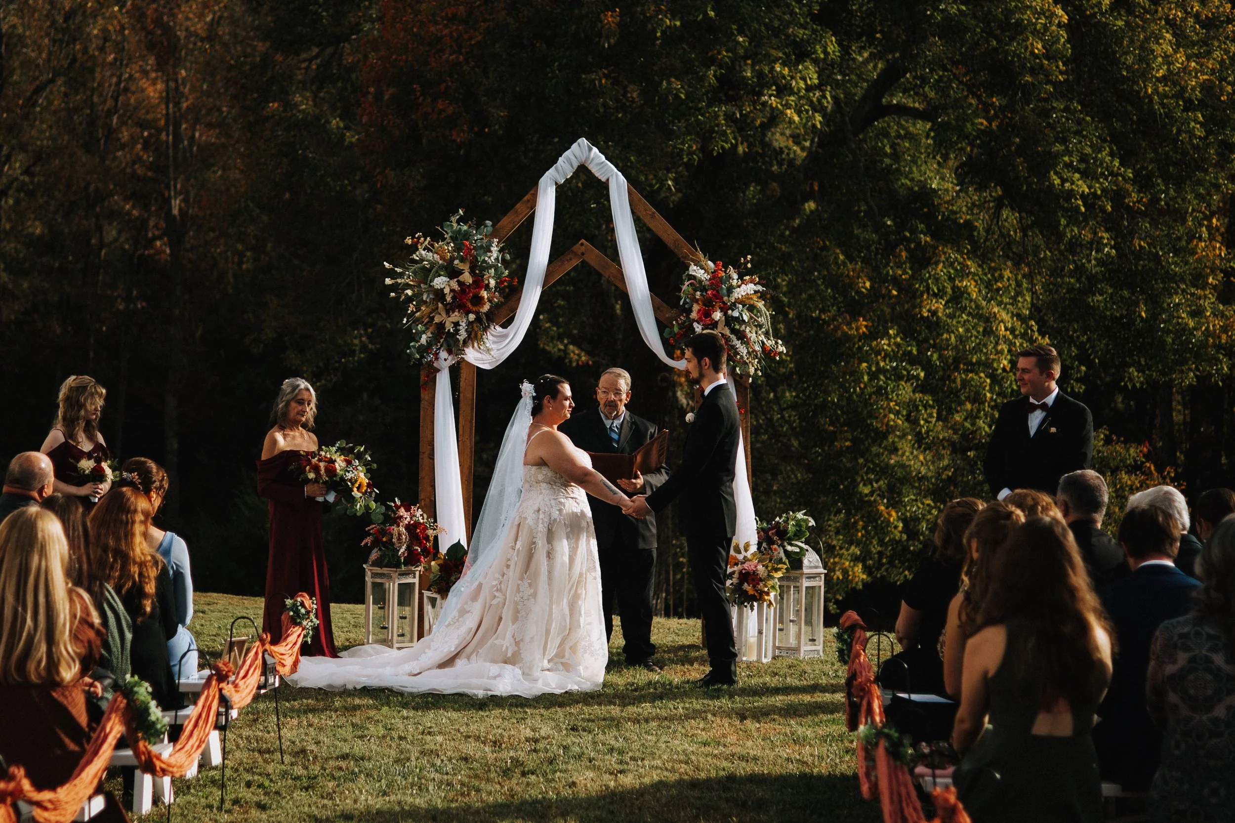 A wedding ceremony outdoors in a wooded area with a small wooden arch decorated with white fabric and flowers, where a bride and groom are exchanging vows, surrounded by seated guests.